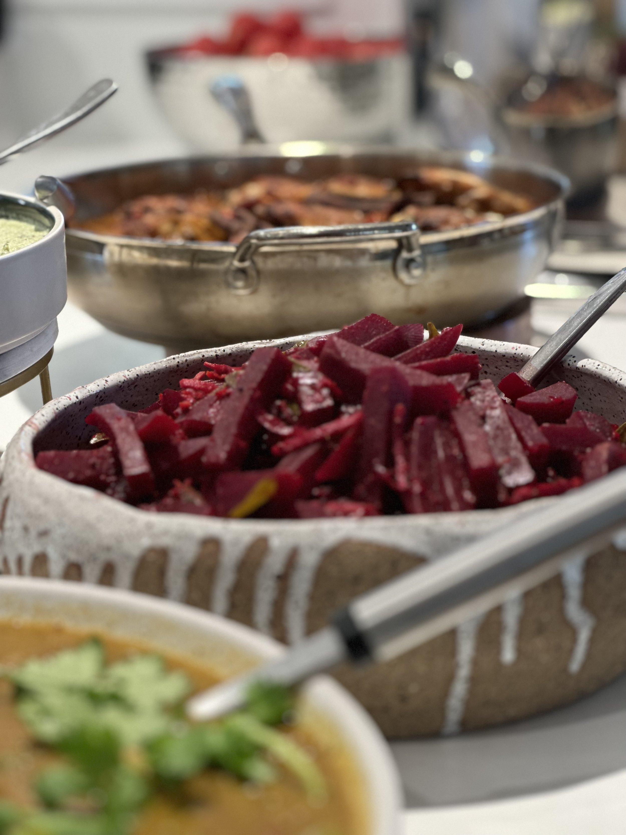 A close-up of a buffet spread featuring a bowl filled with sliced beets, another dish with herb garnish, and a metal serving dish in the background.