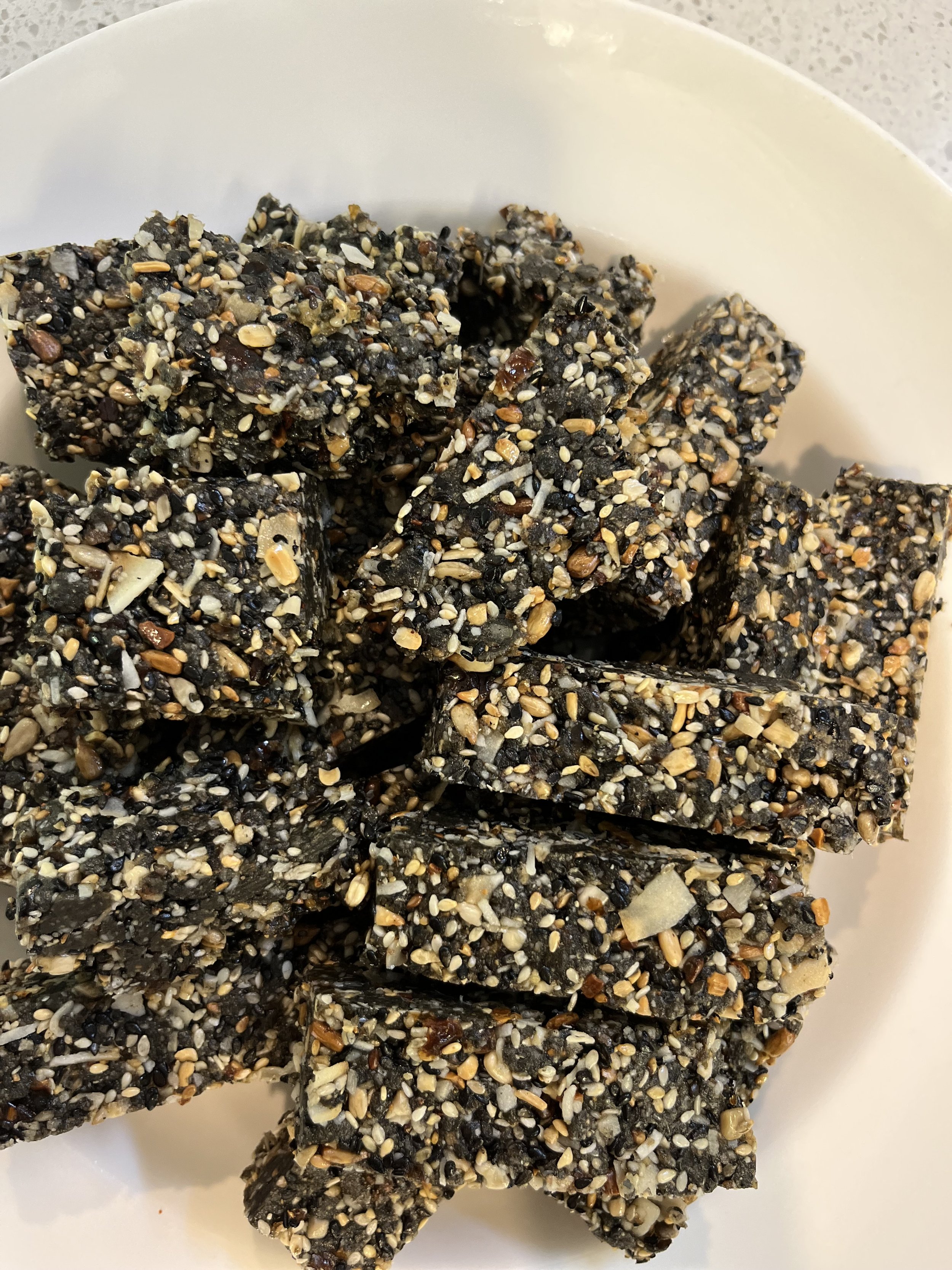 A close-up of seed bars on a white plate, featuring a mixture of black sesame seeds, white sesame seeds, and assorted grains.