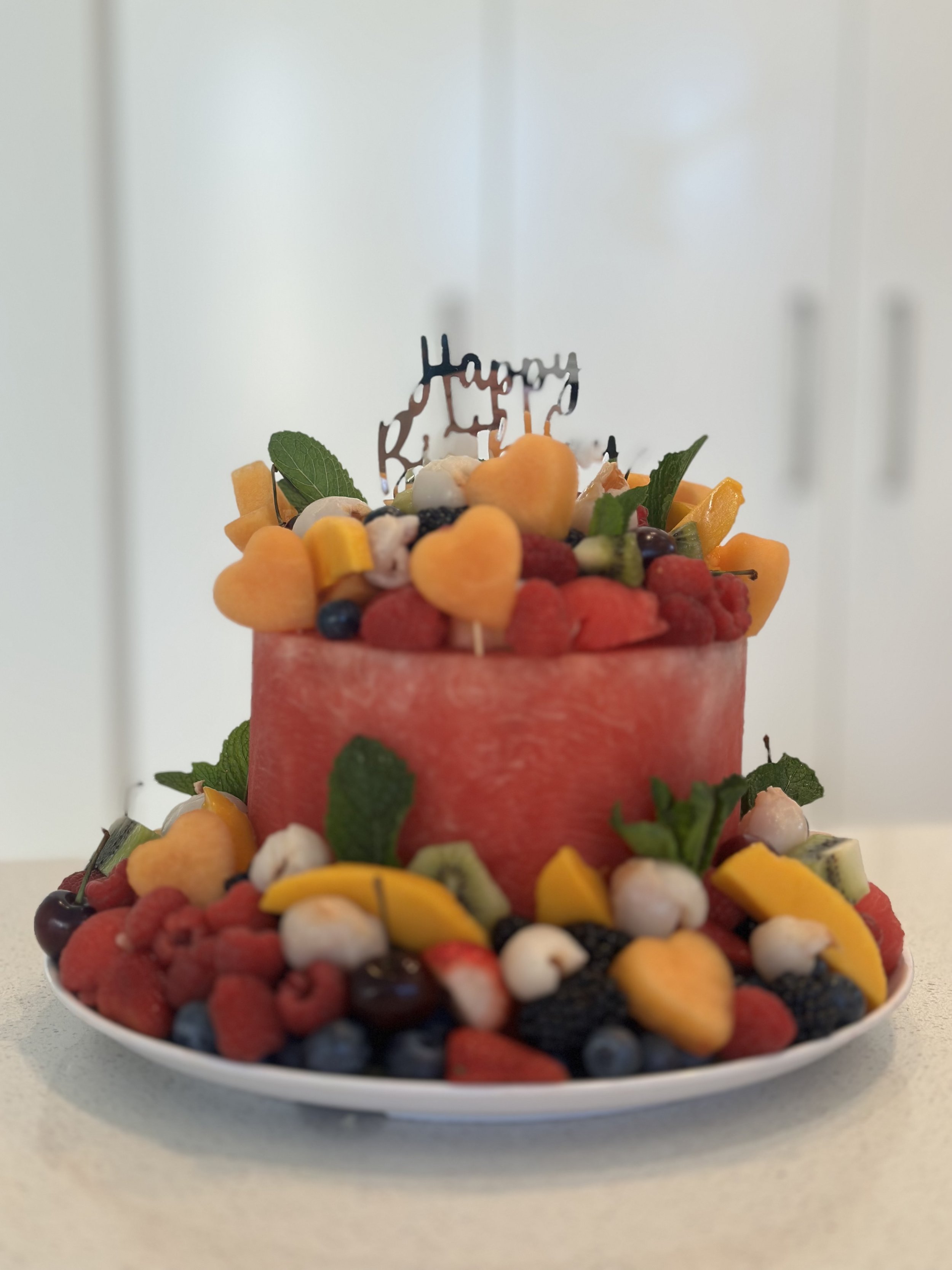 Fruit cake made from watermelon with assorted fruits and berries on a plate, decorated with a "Happy Birthday" topper.