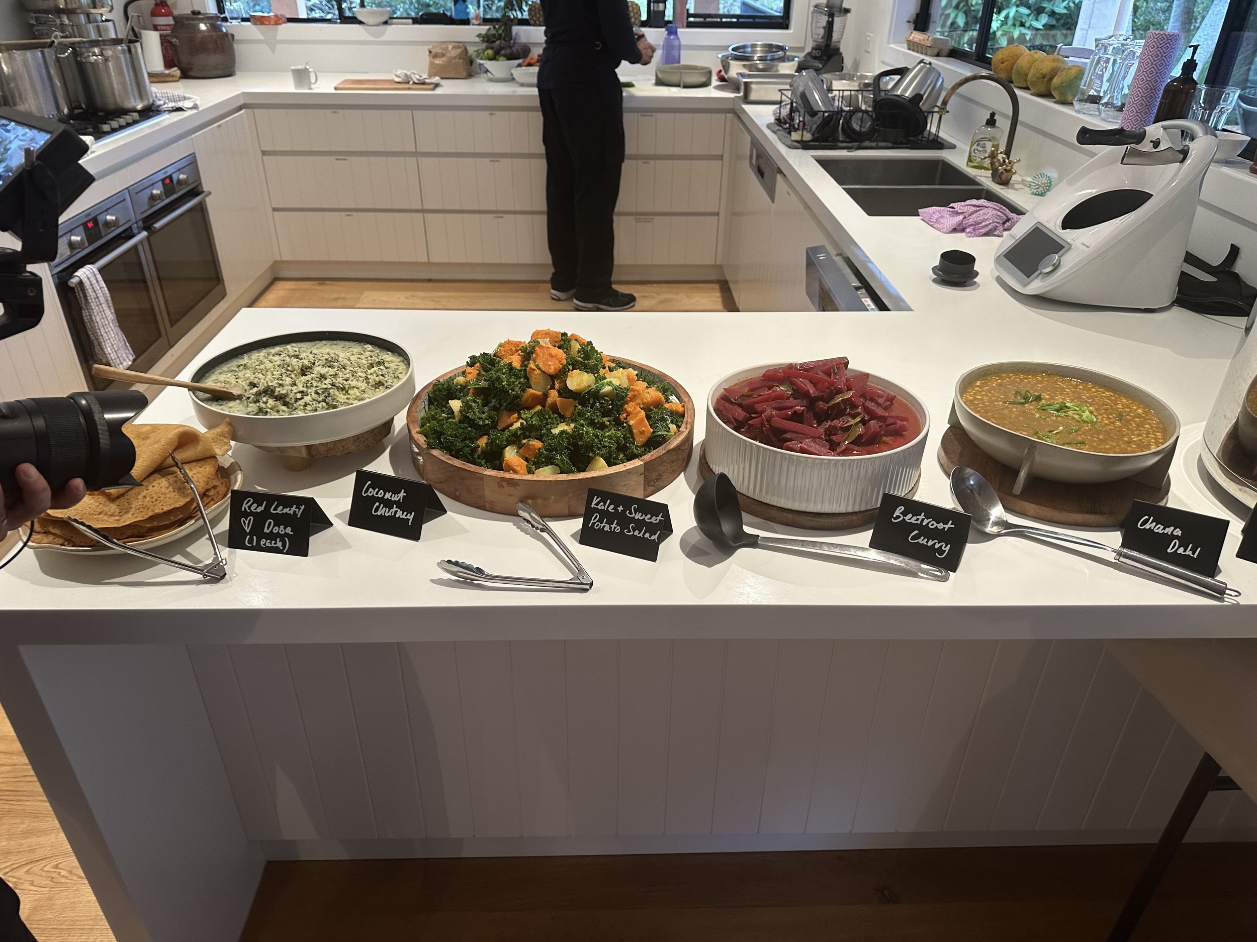 Kitchen counter with a variety of dishes including red lentil dosa, coconut chutney, kale and sweet potato salad, beetroot curry, and chana dahl.