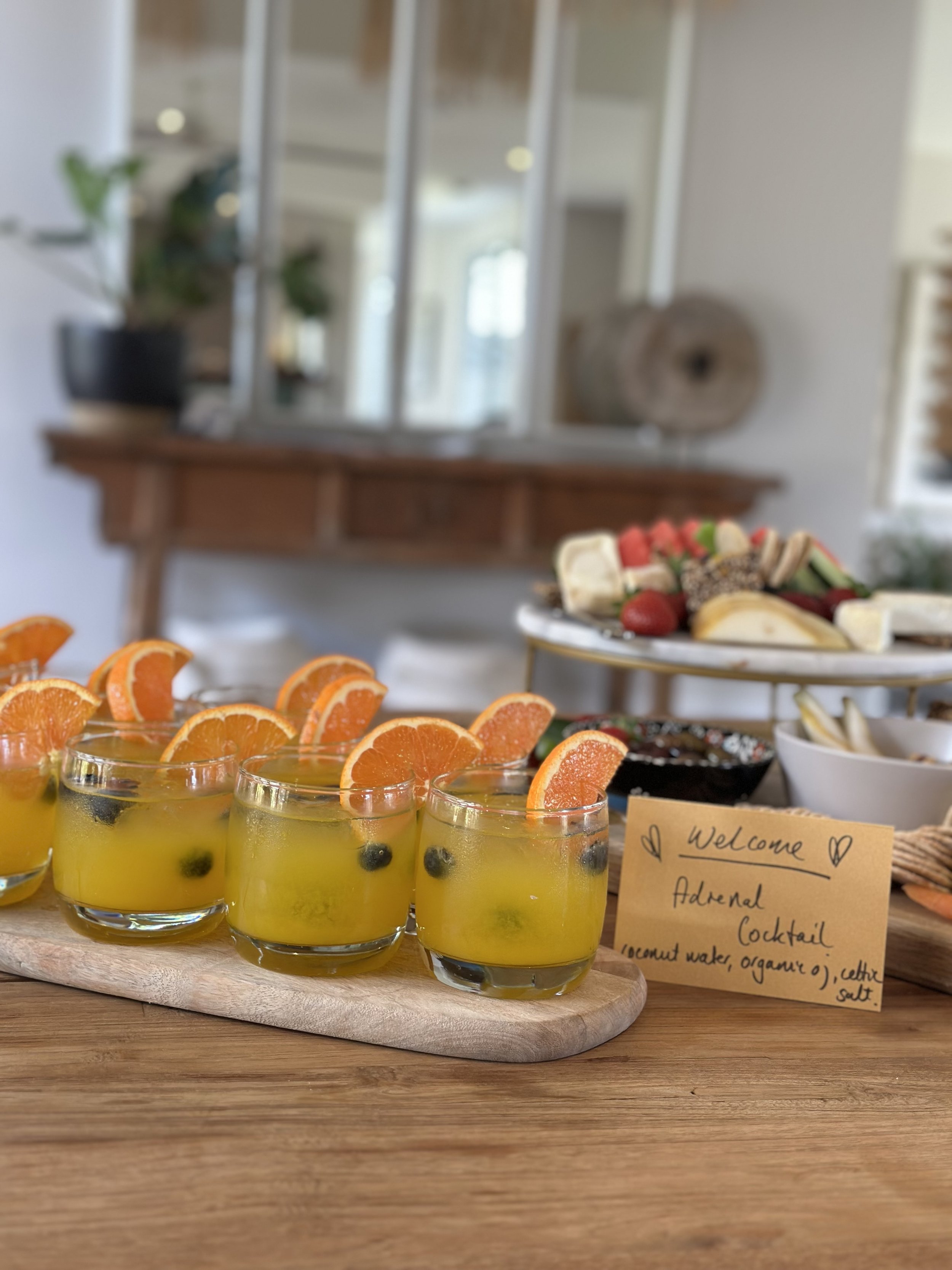 Orange juice cocktails with sliced oranges on a wooden board, accompanied by a sign reading "Welcome Adrenal Cocktail" with ingredients listed, and a platter of fruit and snacks in the background.