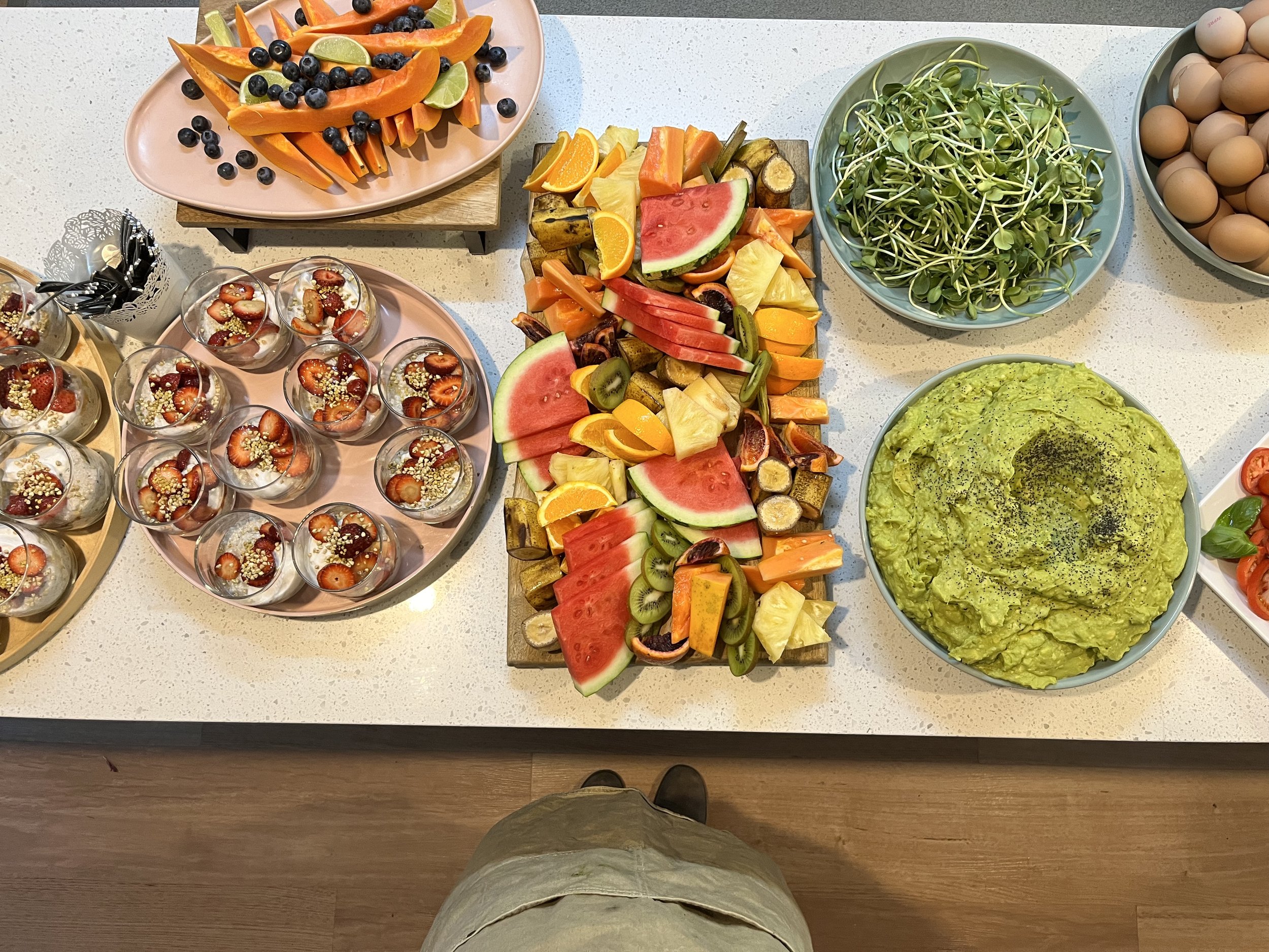 A selection of fruits, salads, and eggs including papaya slices with blueberries and lime wedges, chia pudding with strawberries, mixed fruit platter, sunflower sprouts, a bowl of guacamole, and a plate of brown eggs on a countertop.