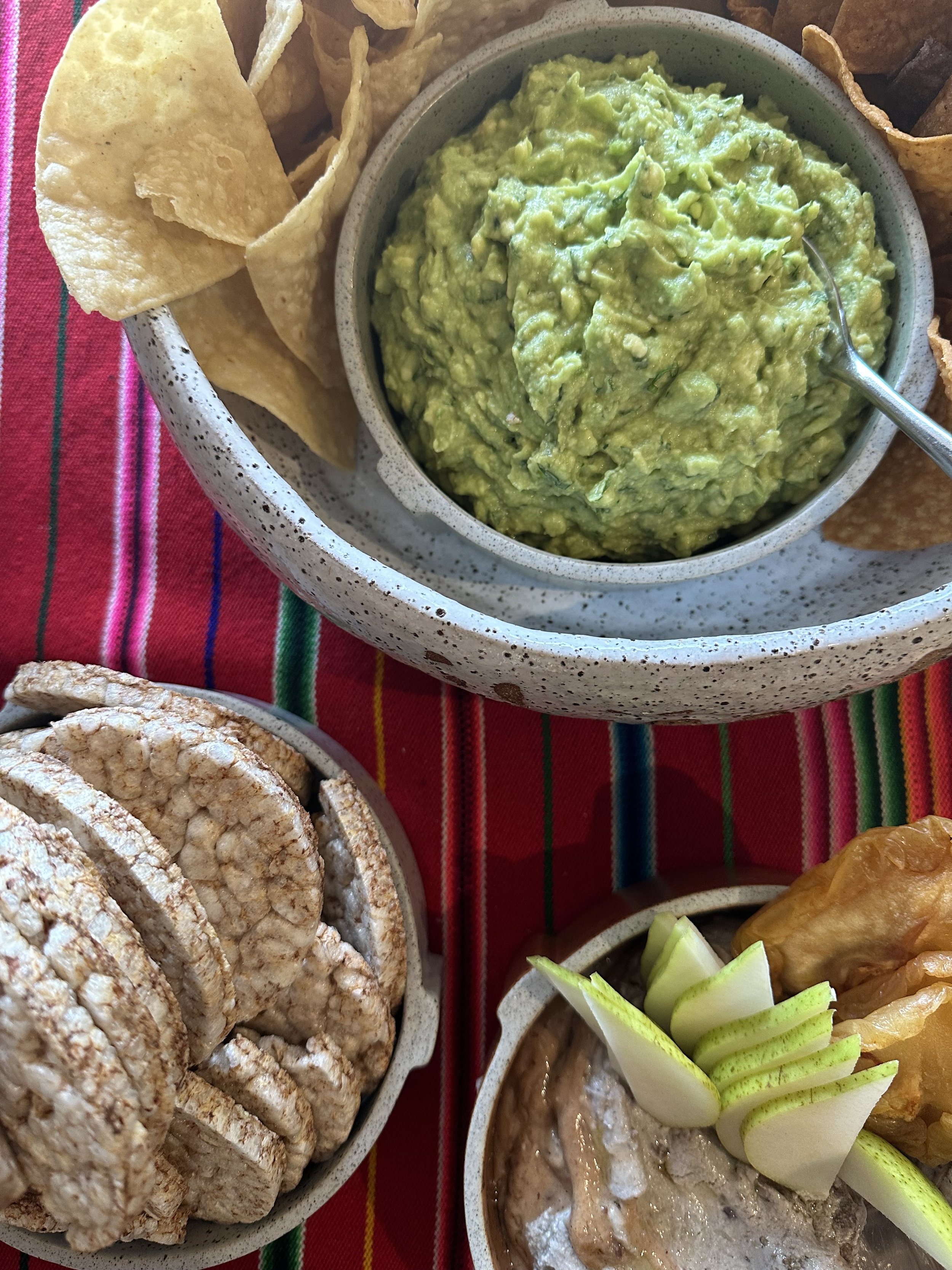 A table setting featuring a bowl of guacamole with tortilla chips, a bowl of rice cakes, and a dip garnished with pear slices and chips on a colorful striped cloth.