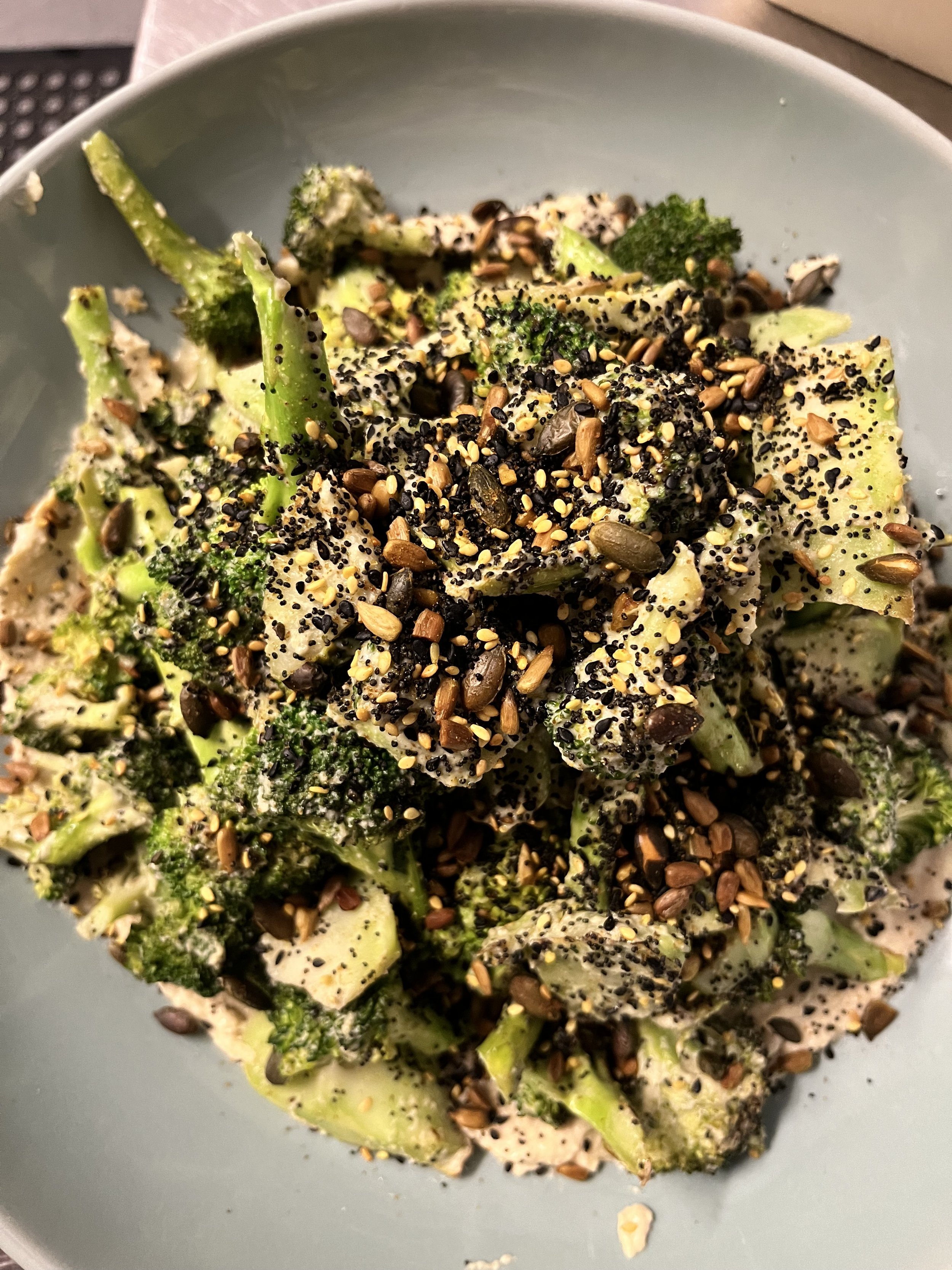 Bowl of broccoli salad topped with seeds on a light gray plate.