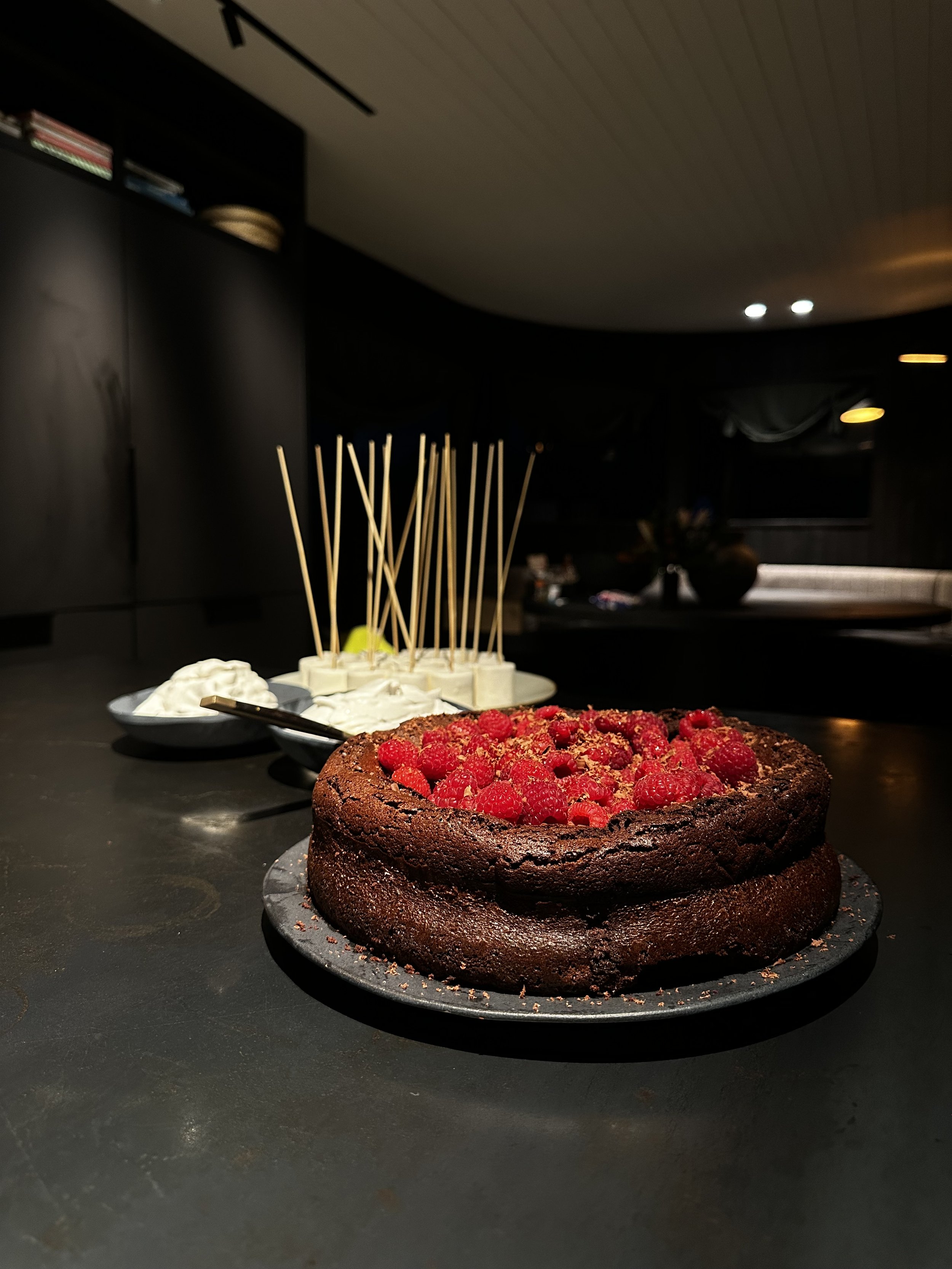 Chocolate cake topped with raspberries on a dark counter, with bowls of whipped cream and skewers in the background.