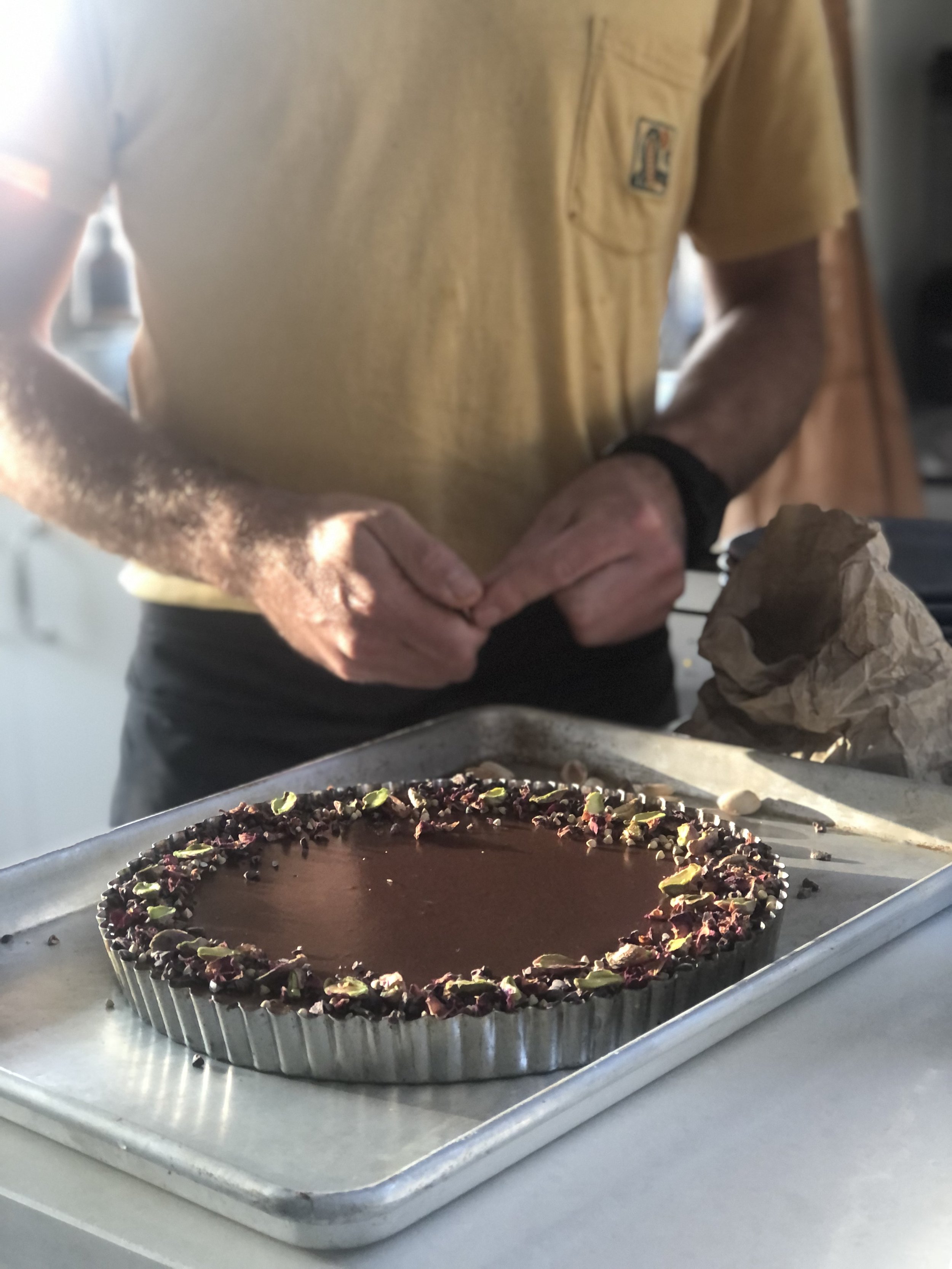 Person in yellow shirt preparing chocolate tart on a metal tray, decorated with nuts and dried fruits in a kitchen setting.