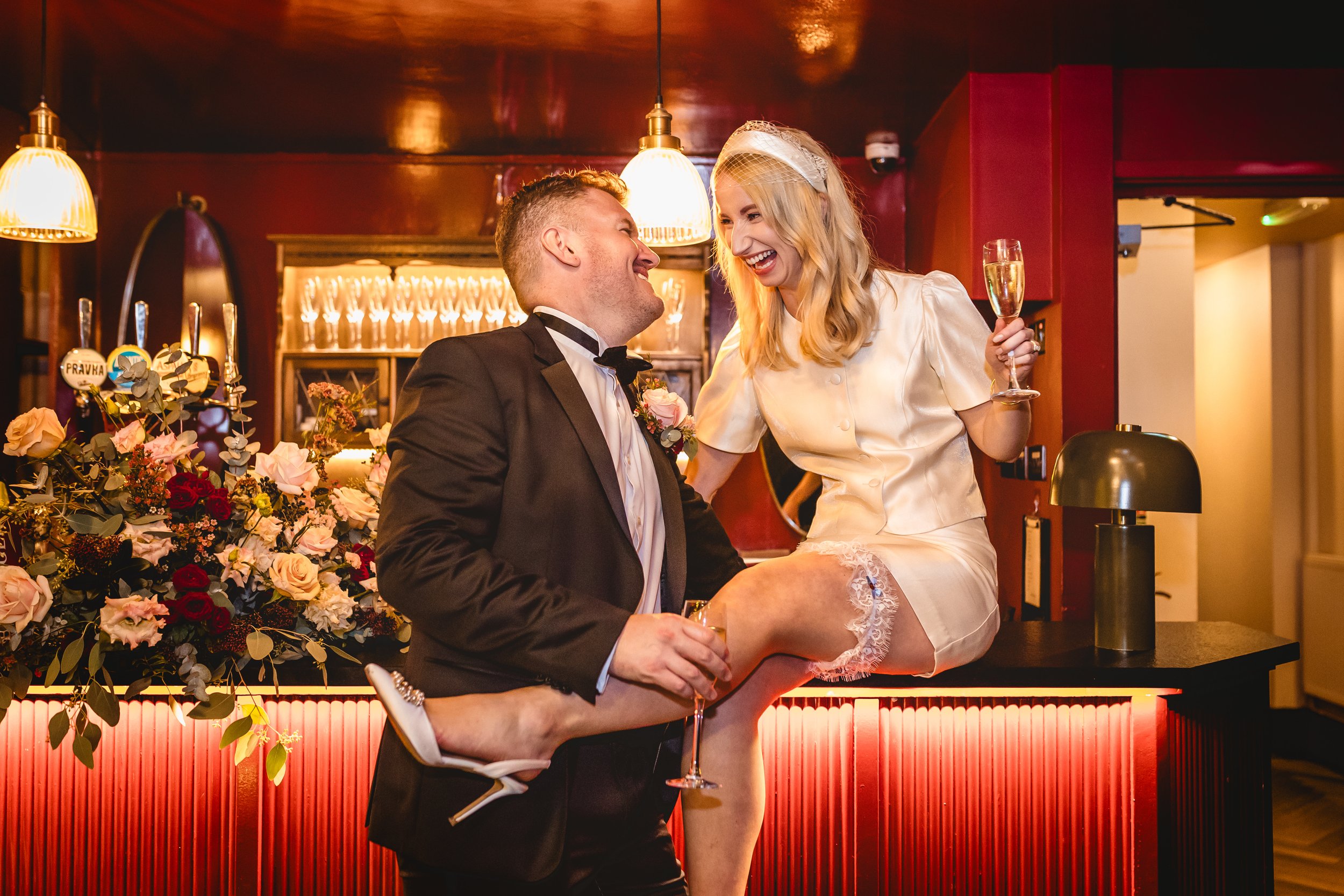 A bride and groom pose for a photo on their wedding day in a stylish, contemporary bar area of a restaurant. The bride sits upon the bar candidly facing her groom as drink Champange and smile.