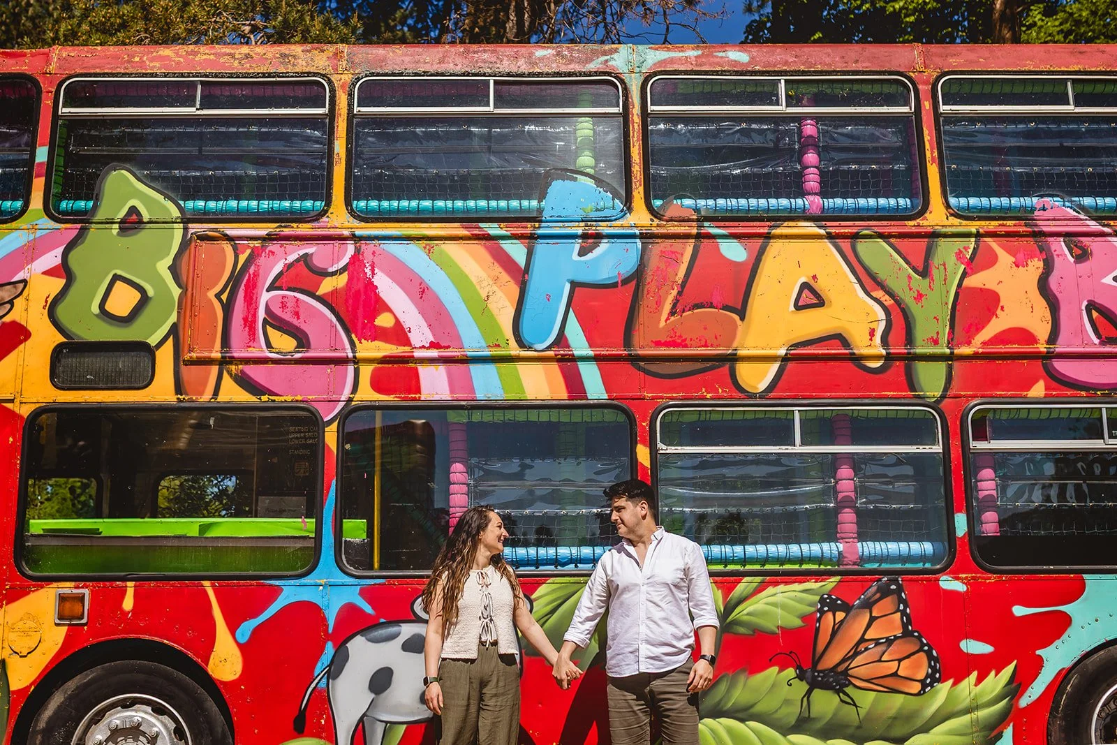 A MAN AND WOMAN STAND IN FRONT OF A RED DOUBLE DECKER BUS THAT IS COVERED IN PLAYFUL AND COLOURFUL GRAFFITTI. THEY HOLD HANDS AND ARE LOOKING TOWARDS EACH OTHER