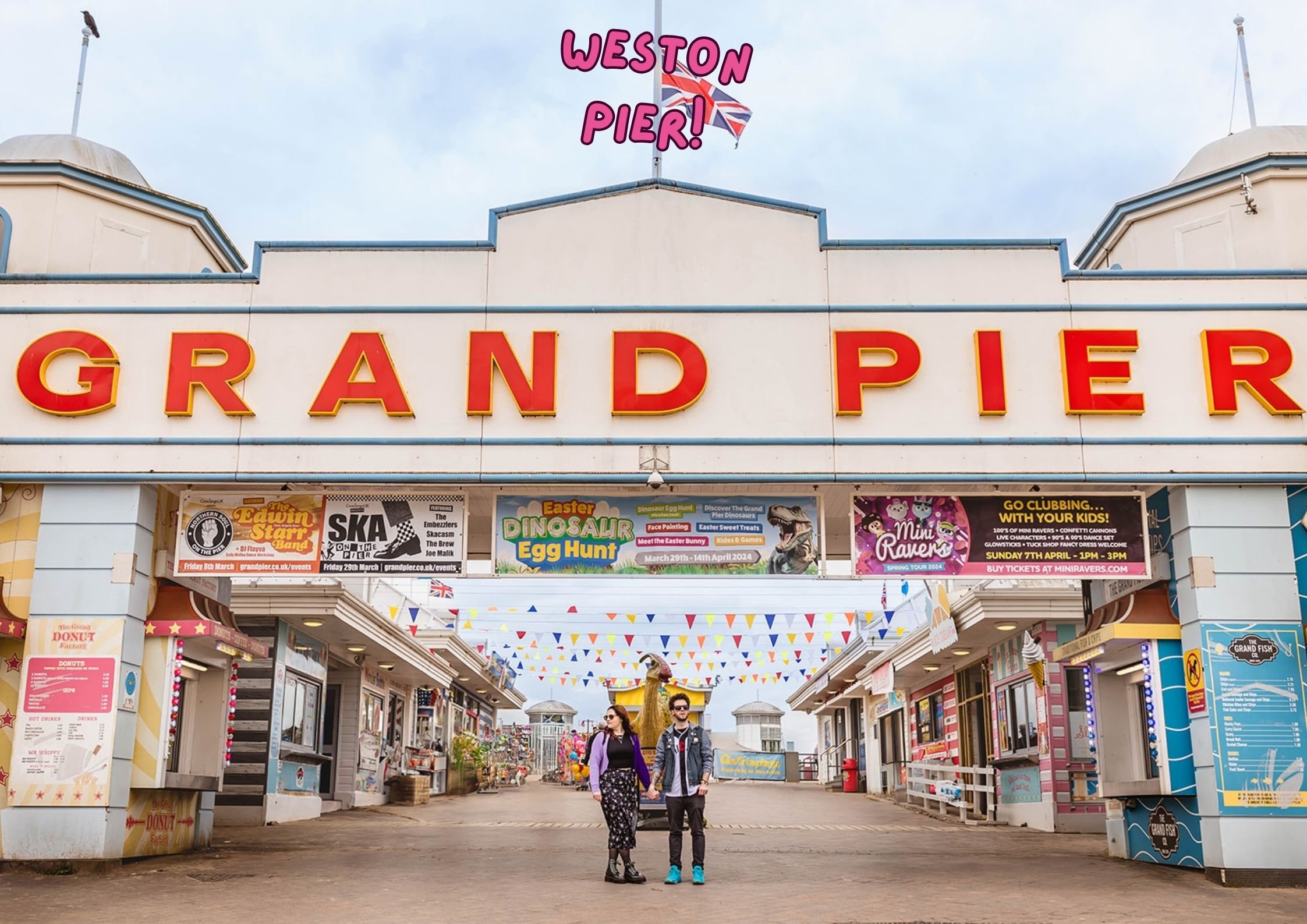 A MAN AND WOMAN POSE WHILST HOLDING HANDS AND LOOKING CANDIDLY WHILST STOOD UNDERNEATH THE LARGE SIGN OF WESTON SUPER MARE PIER