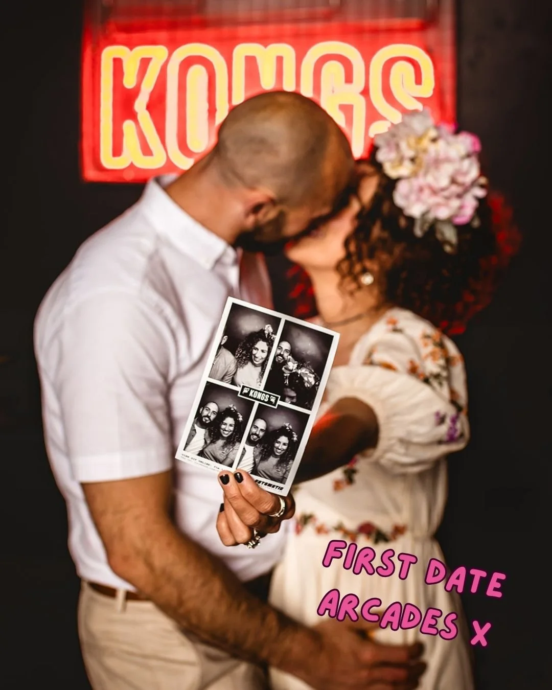 AN OUT OF FOCUS COUPLE KISSING ARE IN VIEW IN THE BACKGROUND WHILST THE WOMAN HOLDS UP A PHOTOBOOTH STYLE PHOTO OF THE TWO OF THEM WHICH IS THE FOCUS OF THE SHOT.