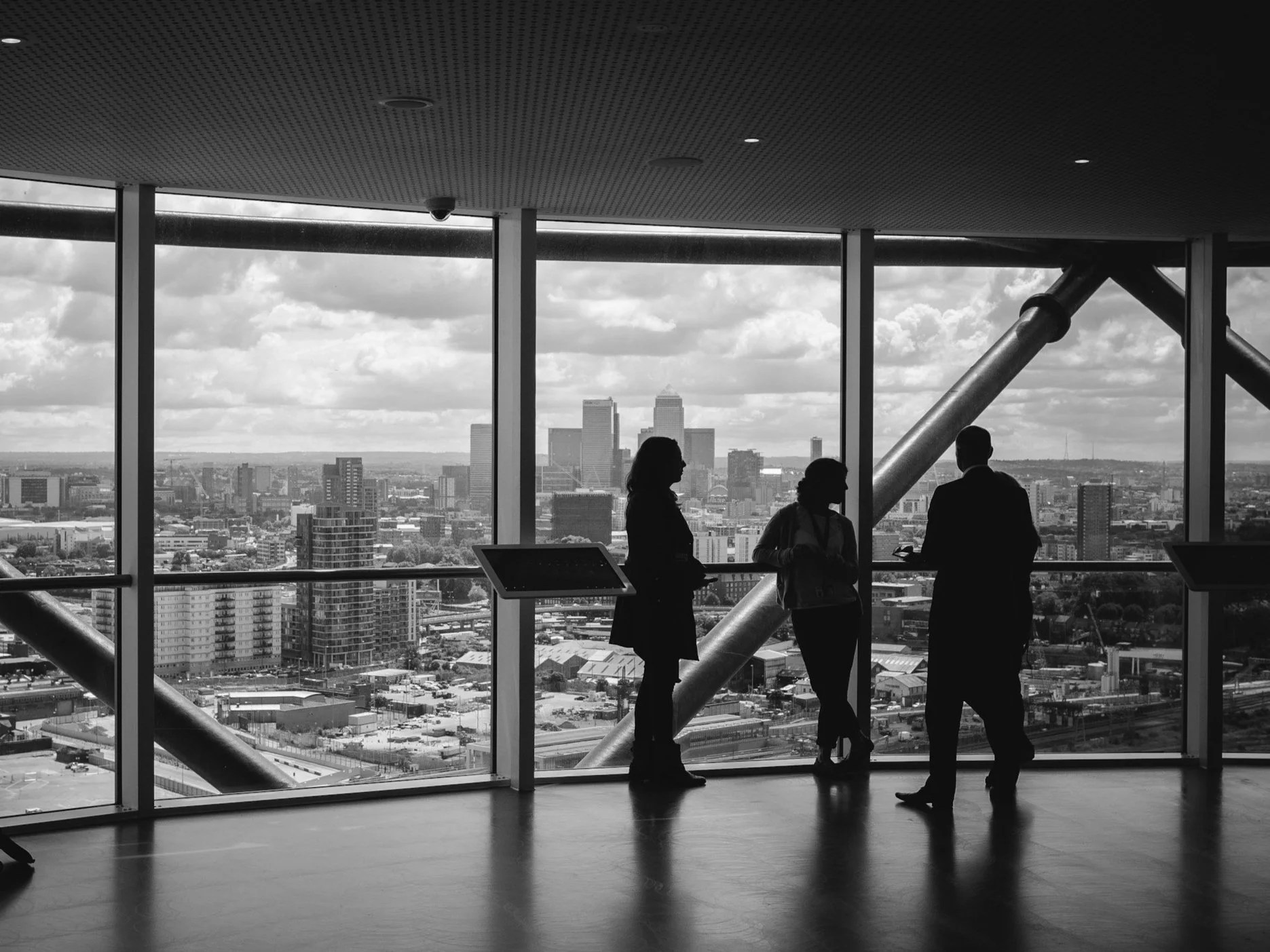 Silhouettes of three people having a conversation in front of large windows with a city skyline in the background during the daytime.
