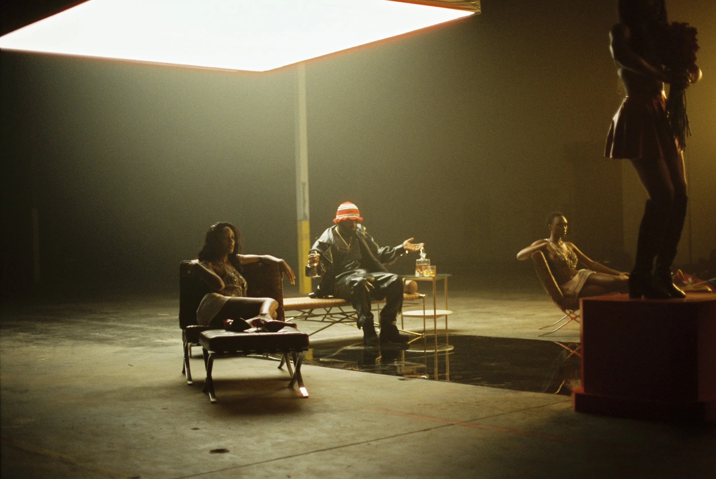 A dimly lit stage with four people, three women sitting on chairs and a man seated in the center, and one woman standing on a platform in the foreground. The scene appears to be part of a theatrical or dance performance.
