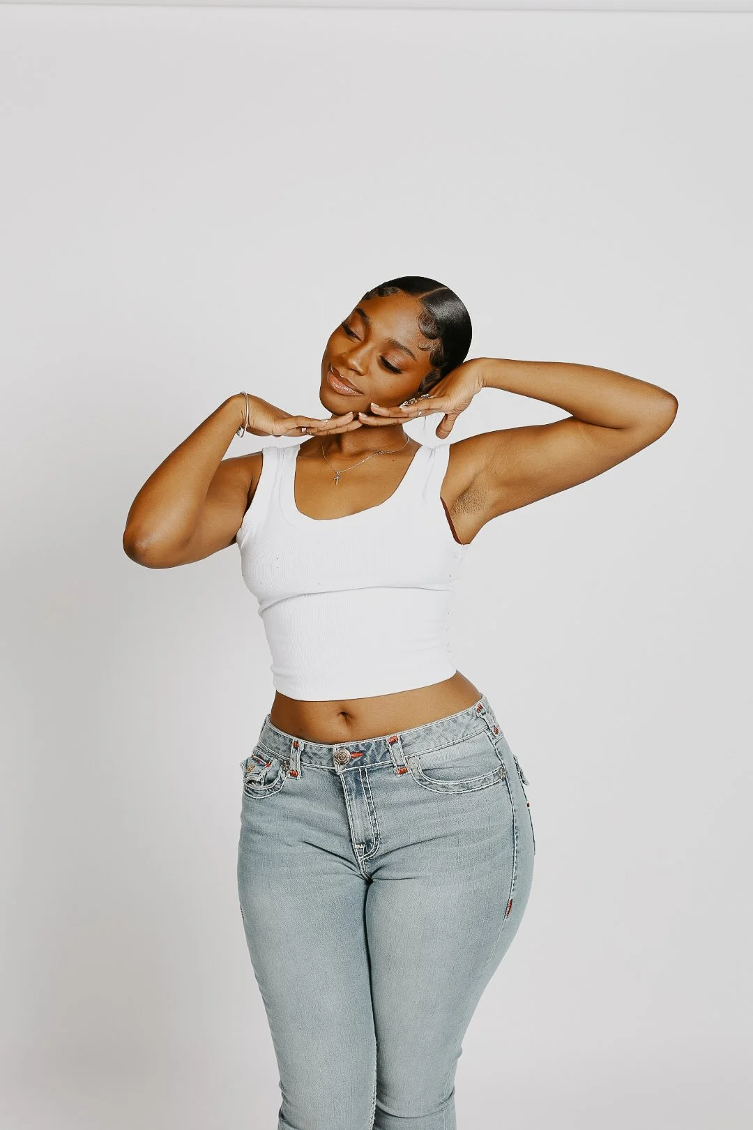 Young woman with short hair in a white tank top and light gray jeans, smiling with eyes closed, posing against a plain white background.