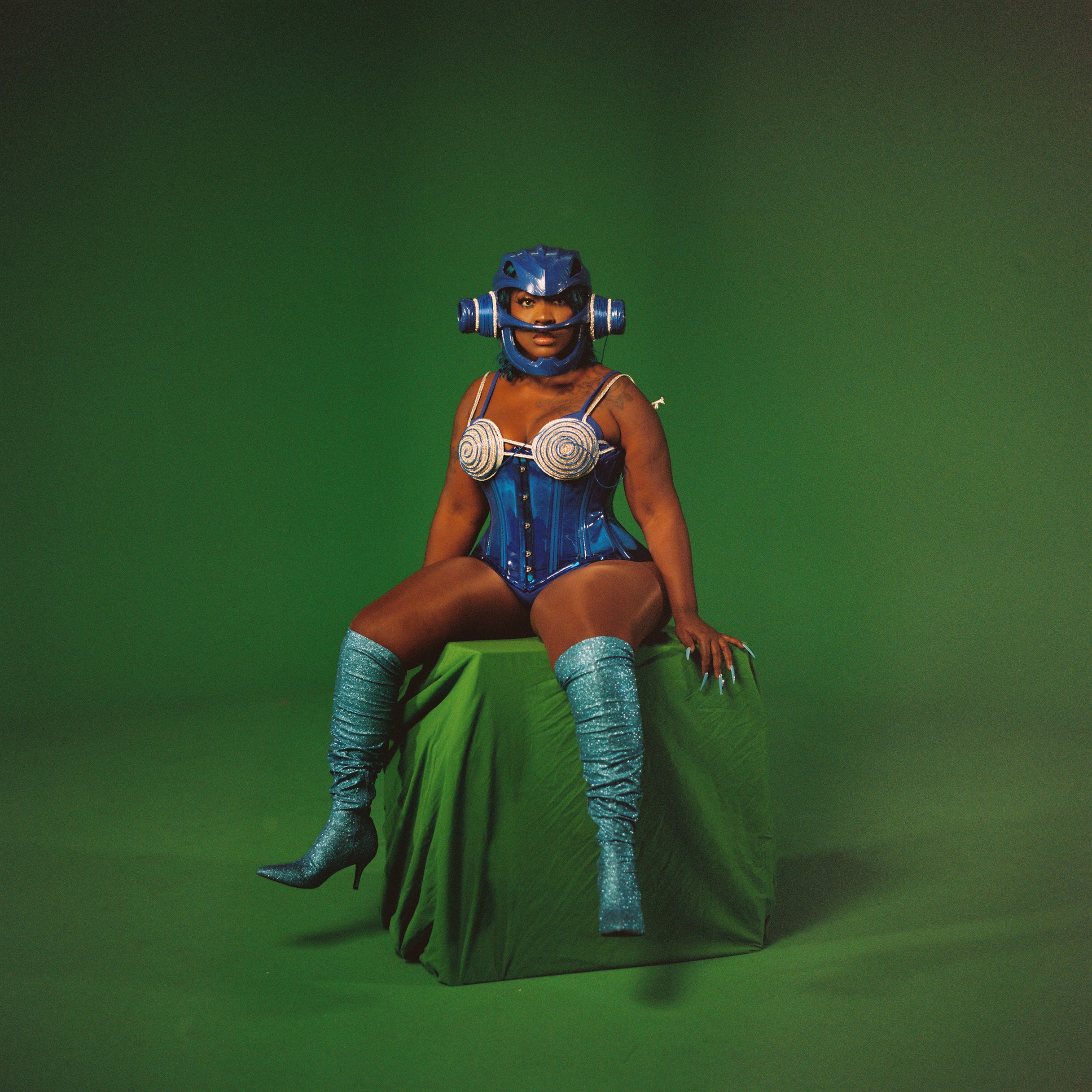 A woman dressed in a shiny blue costume with a futuristic helmet, sitting on a green covering on a green background.