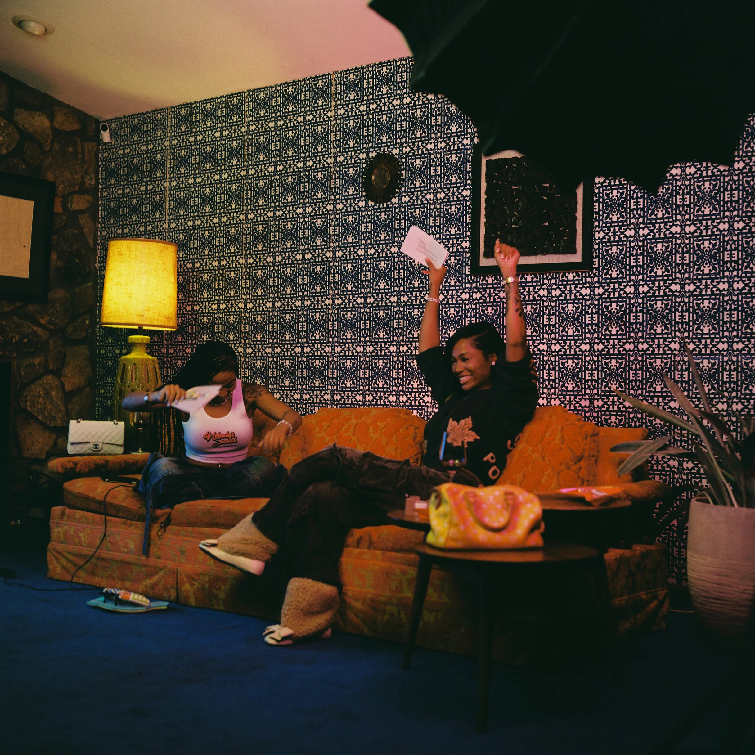 Two women sitting on a vintage orange couch in a cozy room, one smiling and raising her arms, the other looking down at papers, with a yellow lamp, patterned wall, and a small table with a colorful bag.