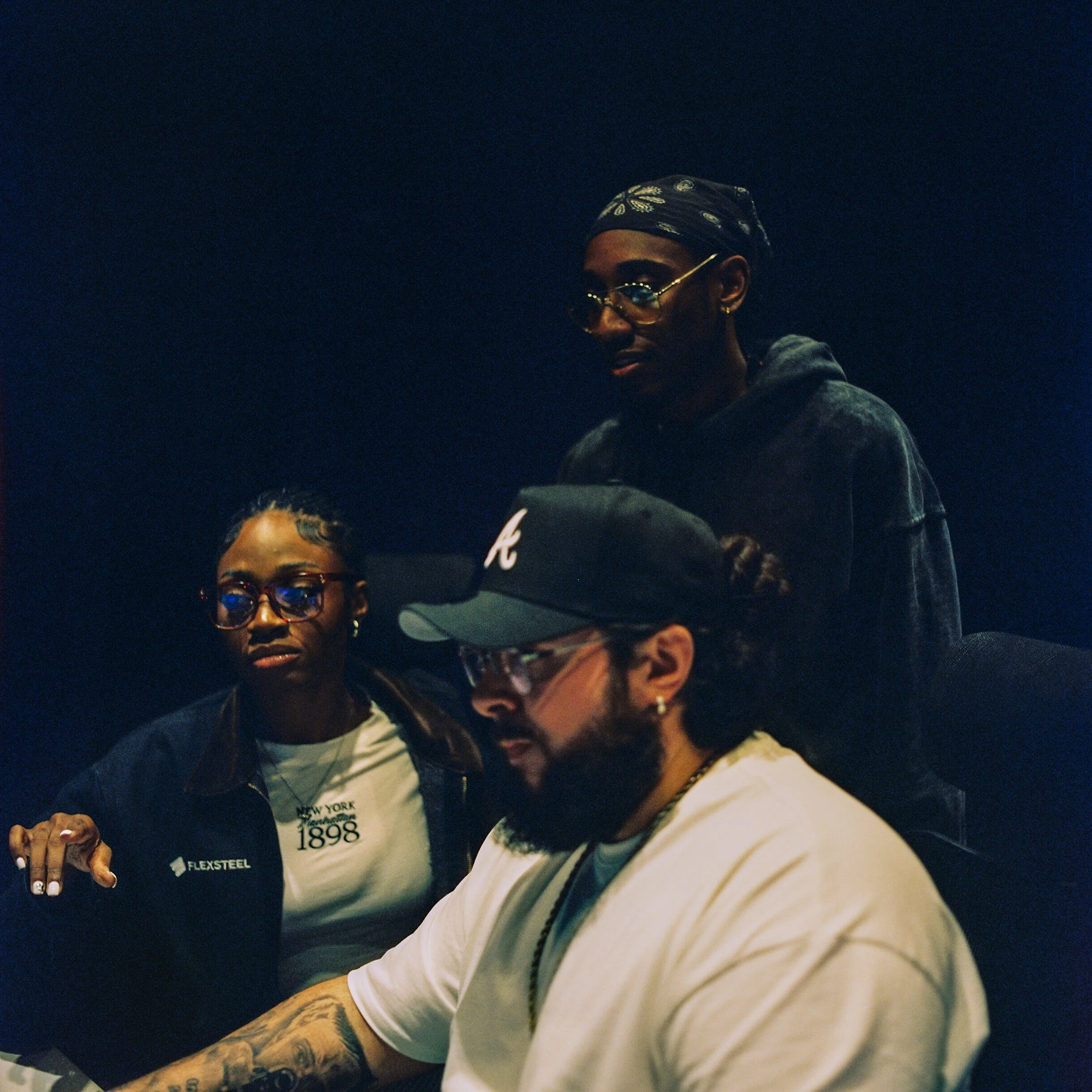 Three people working together in a dark room, focused on a task, with one person wearing a baseball cap and glasses, another wearing richly styled glasses, and a third with a bandana and glasses.