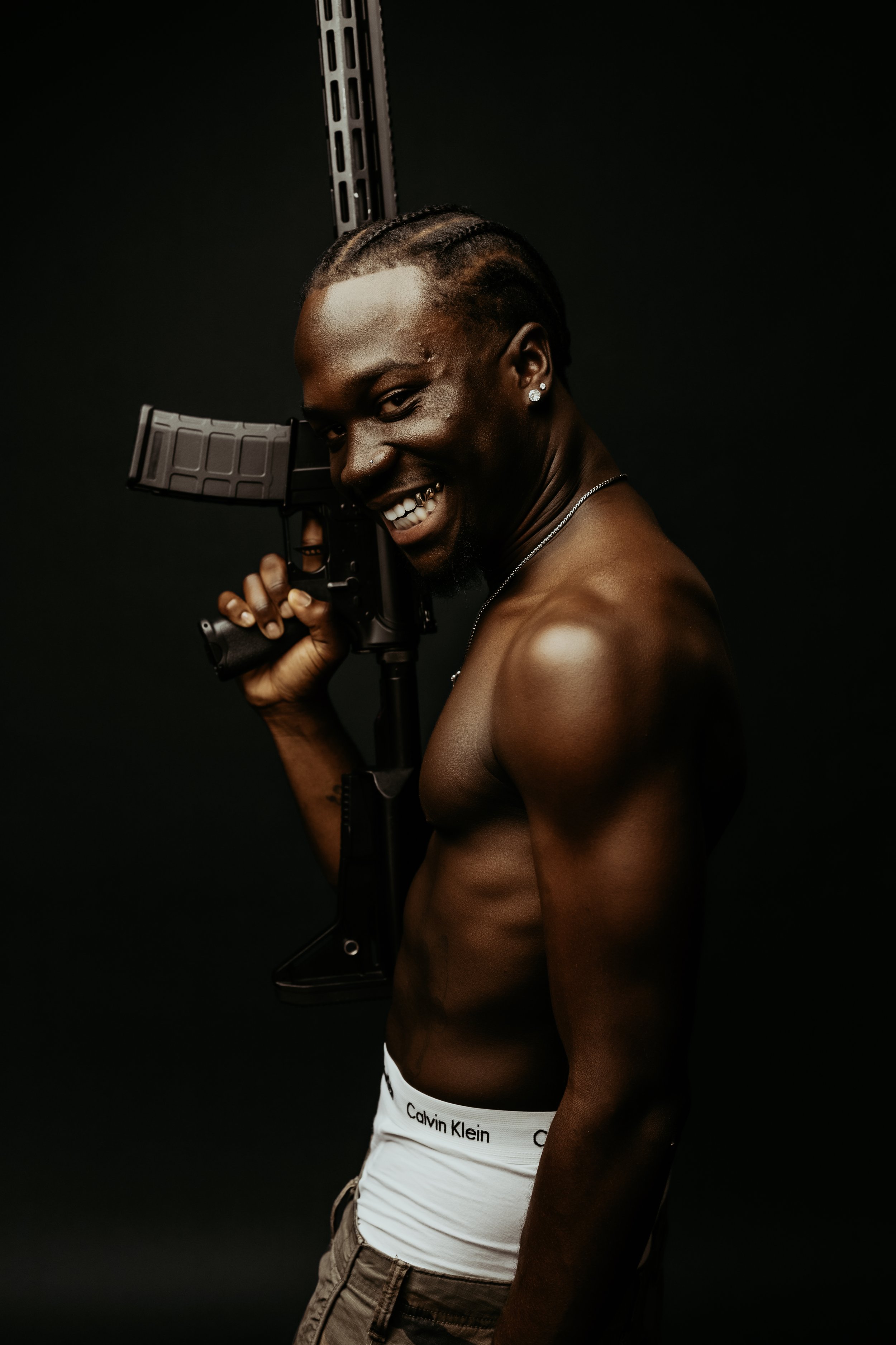 A shirtless young man with cornrows holding a rifle, smiling, against a black background.
