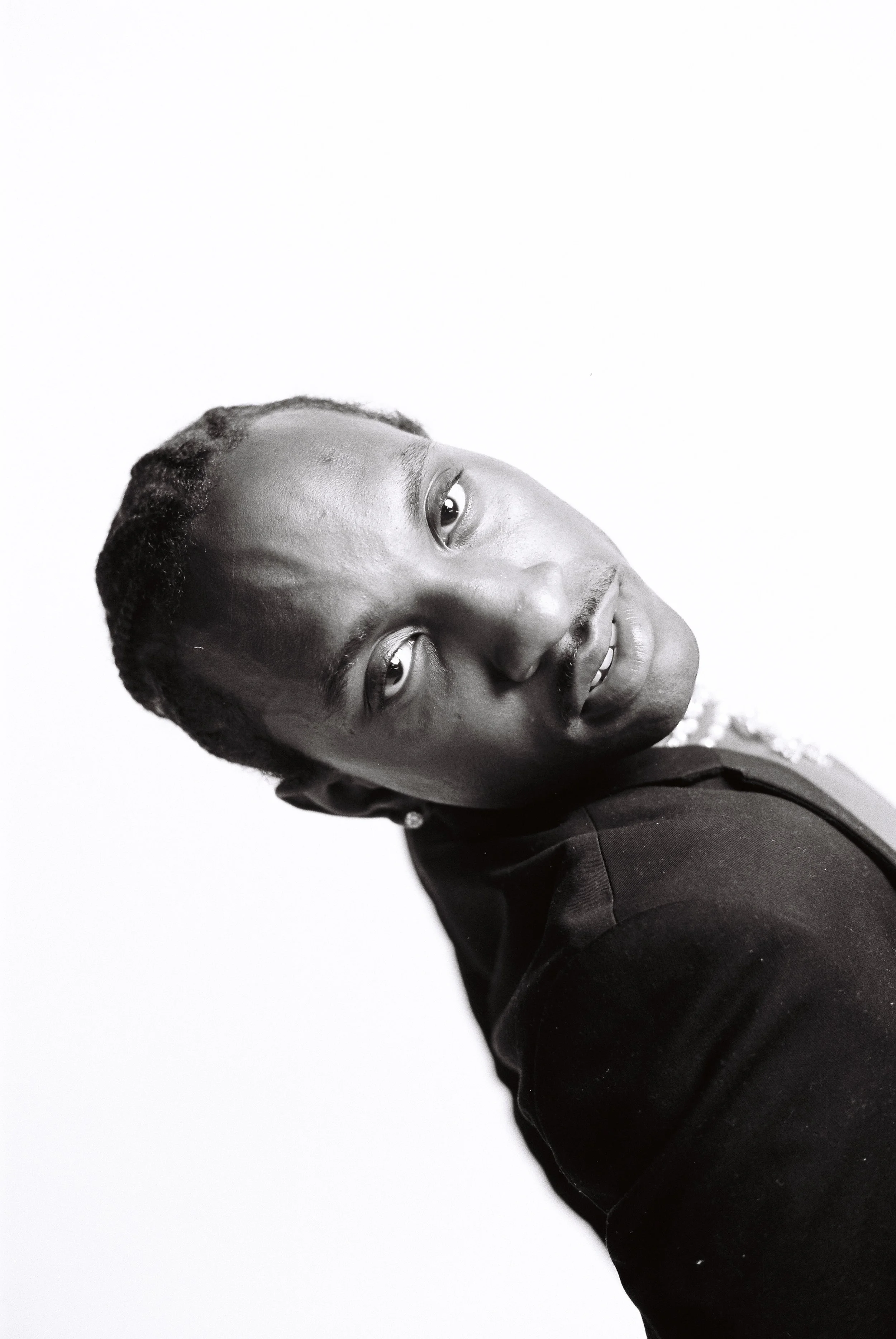 Black and white portrait of a person with short textured hair, wearing a dark outfit, looking directly at the camera with a slight smile, against a plain white background.