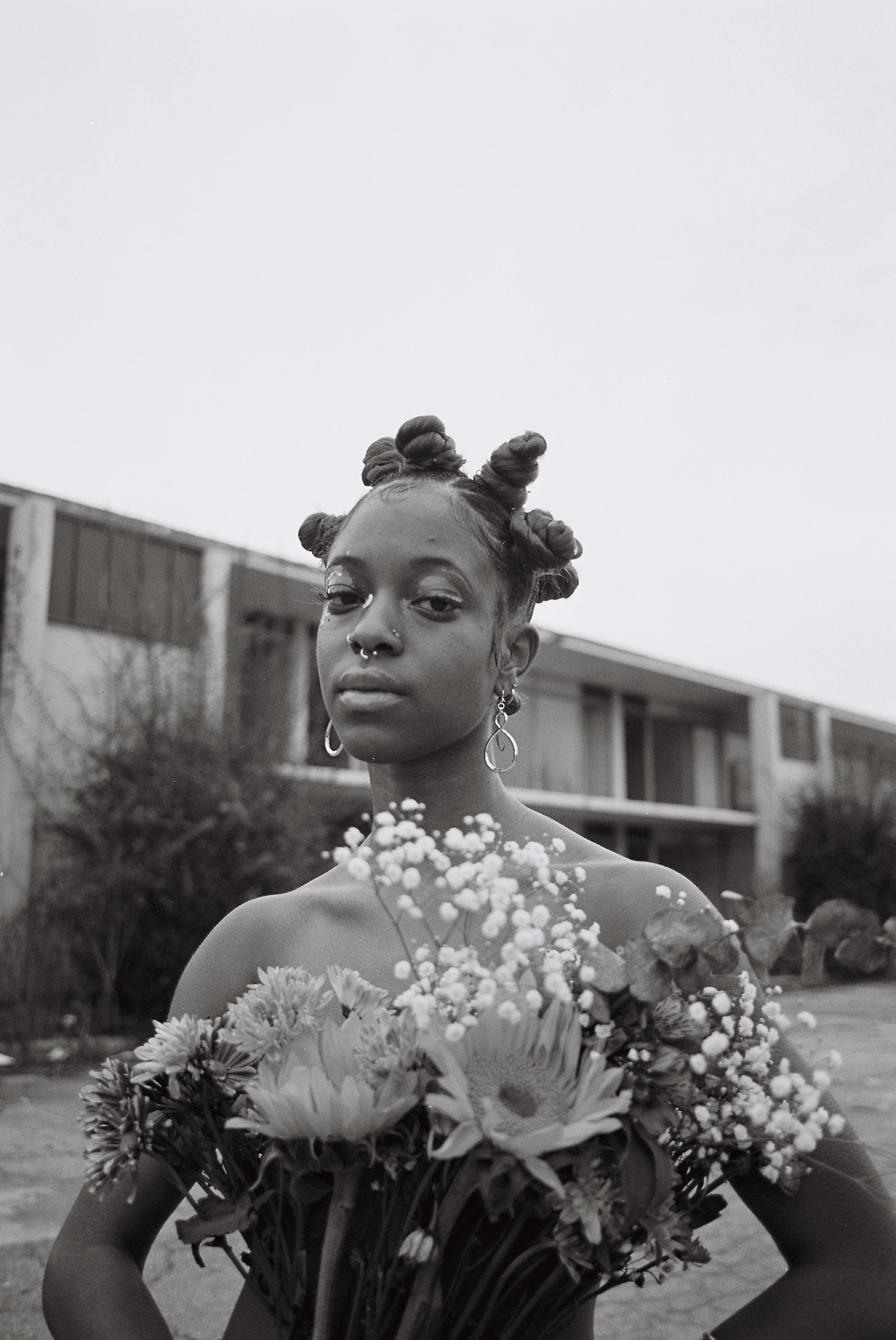 A young woman with styled bantu knots hairstyle holding a bouquet of flowers outdoors in front of a building in black and white.