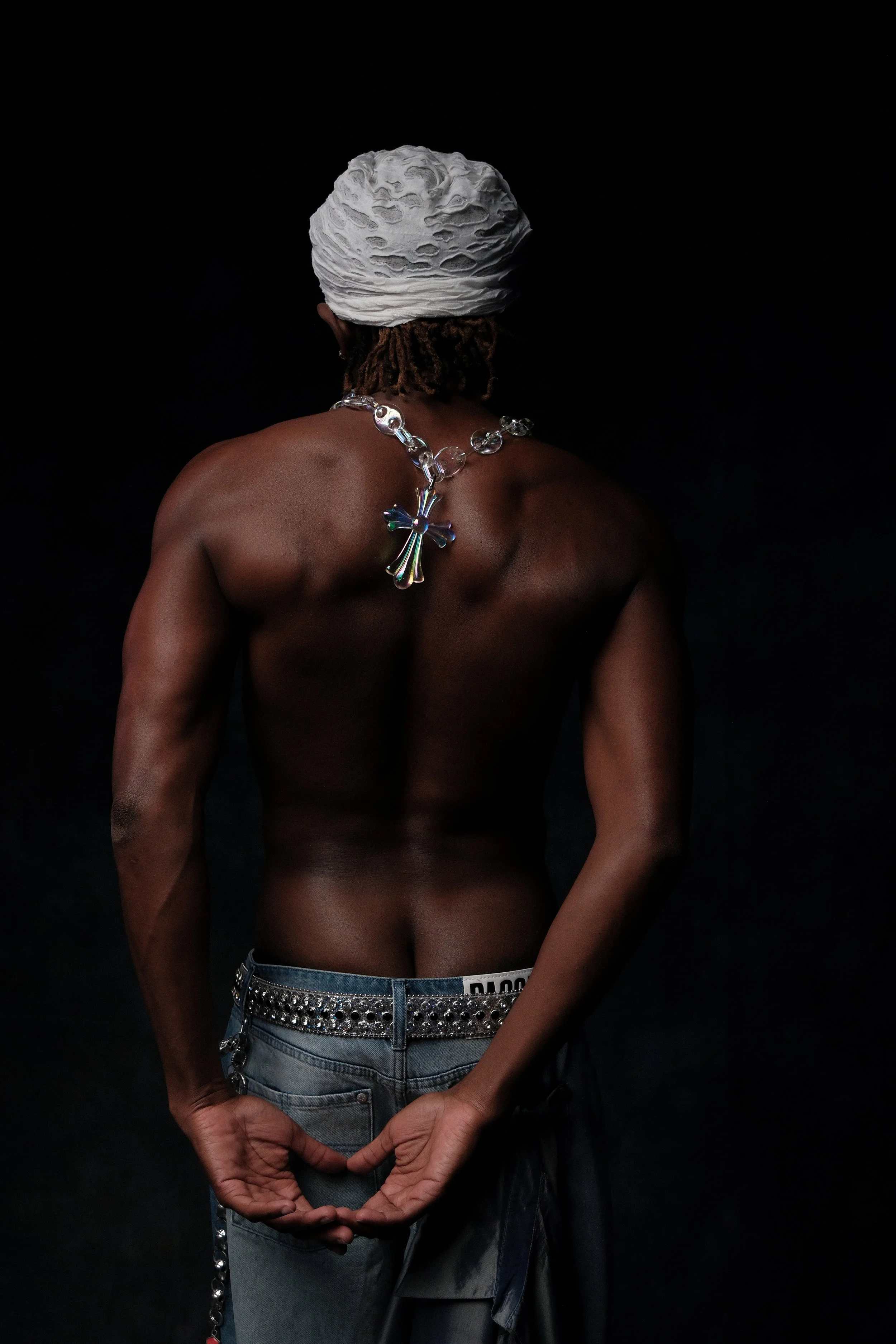 Back view of a shirtless person with dark skin, wearing a white headwrap, a large silver cross necklace, and baggy jeans with a silver studded belt, standing against a dark background.