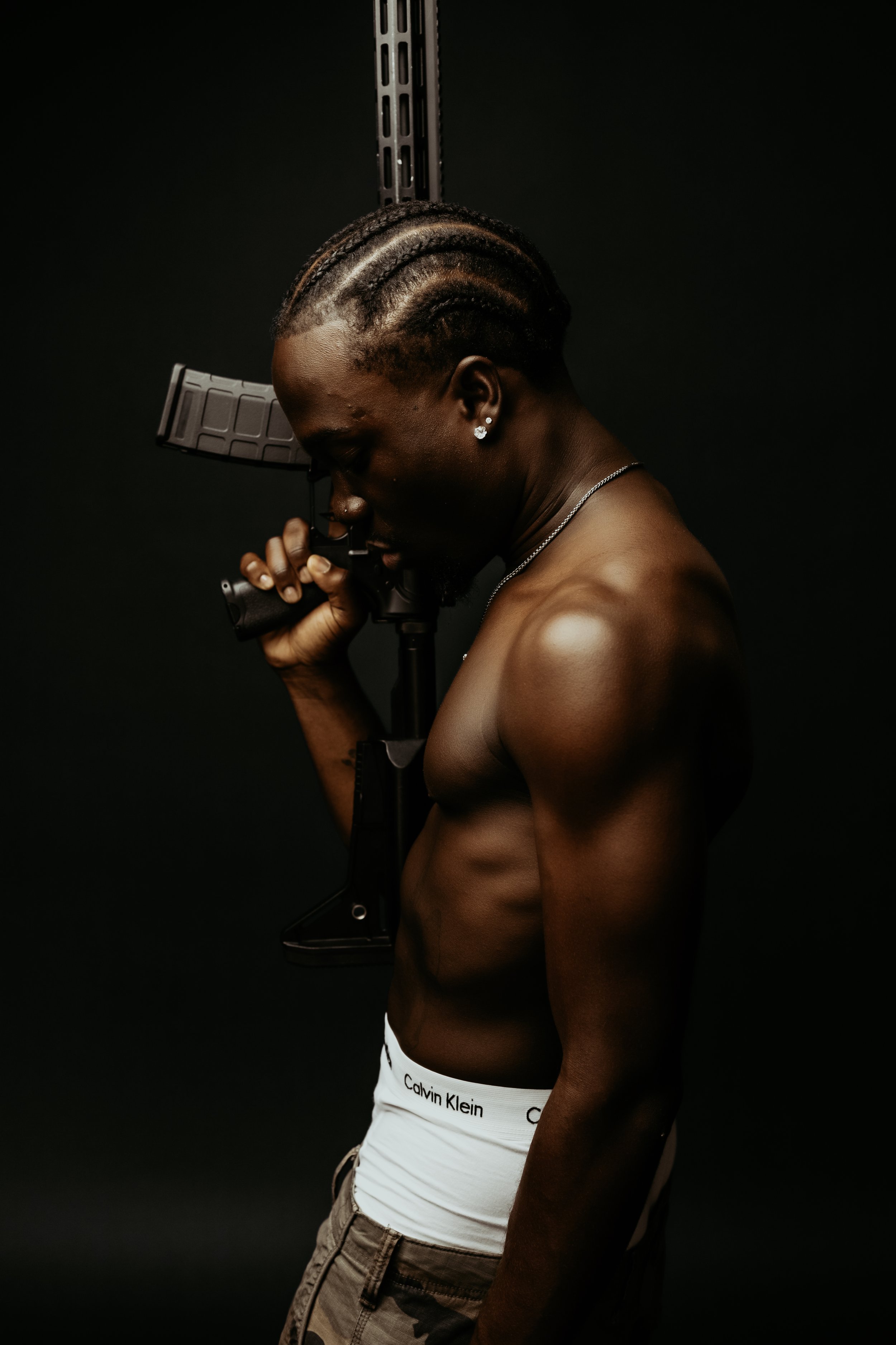 A shirtless man with braided hair and earrings holding a firearm with a dark background.