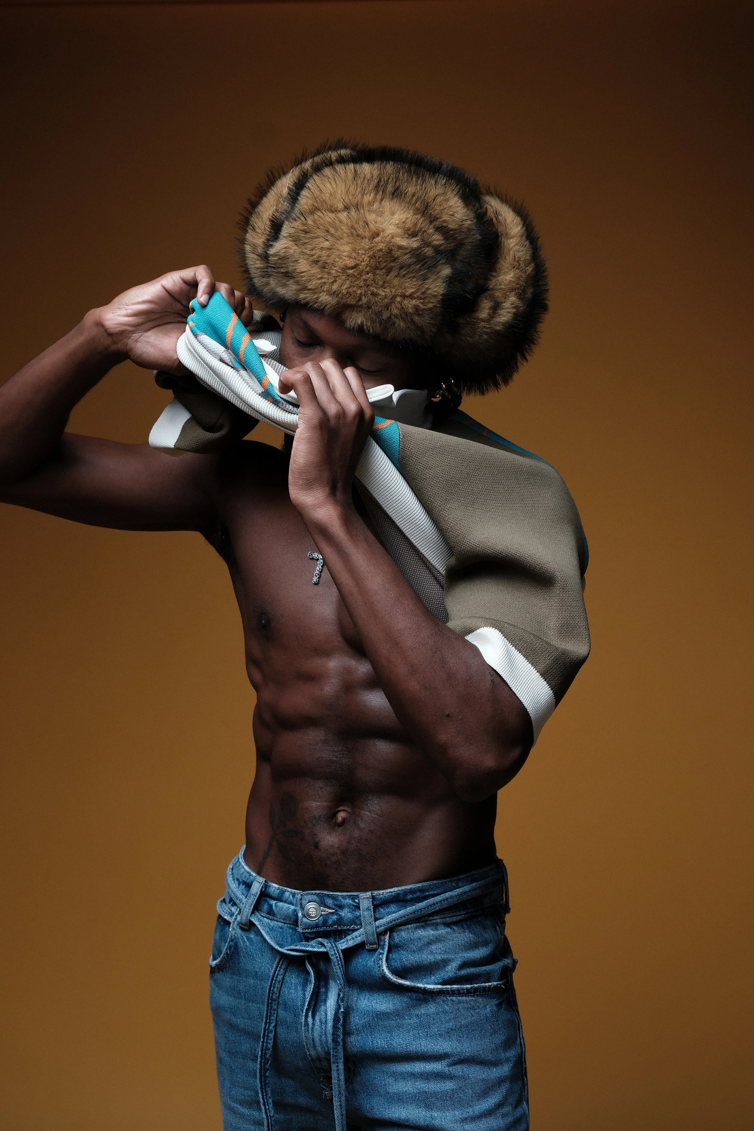 A shirtless man with a muscular physique, wearing blue jeans, a fur hat, and holding a jacket against his face, posing against a plain brown background.