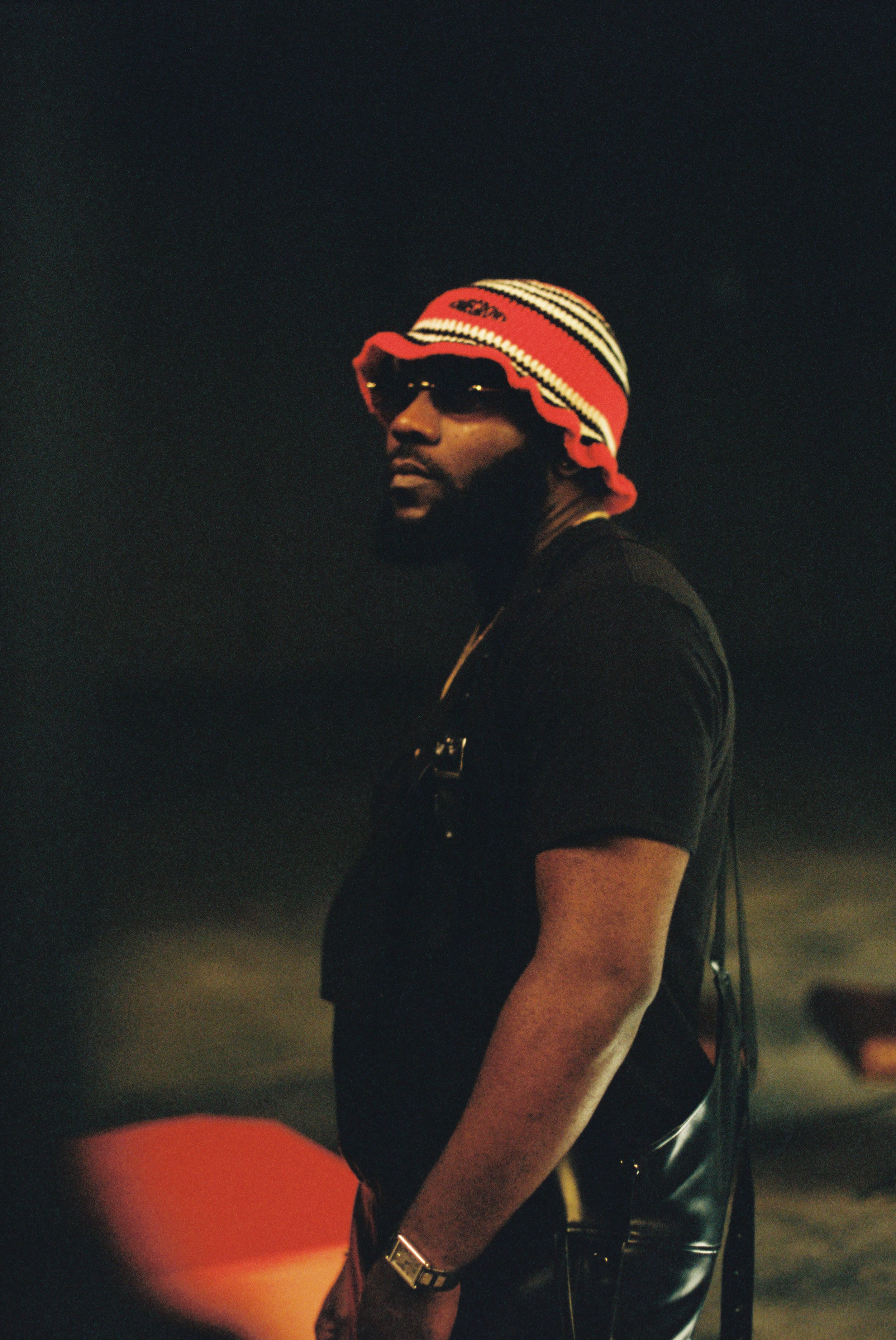 A man with a beard wearing sunglasses, a striped bucket hat, a black t-shirt, and a wristwatch, standing outdoors at night.