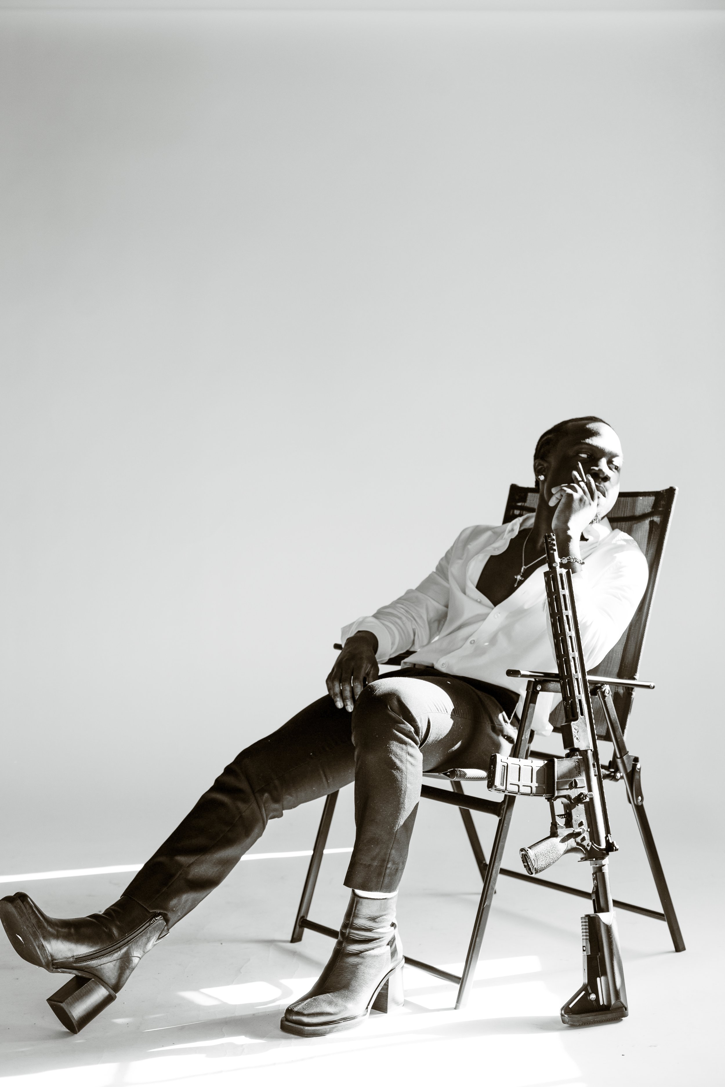 A woman sitting on a chair with a firearm beside her, dressed in a white shirt, dark pants, and boots, in a monochrome studio setting.