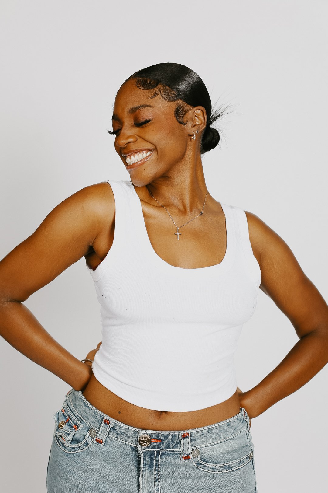 Smiling woman with dark hair in a bun, wearing a white tank top and jeans, standing against a plain white background.