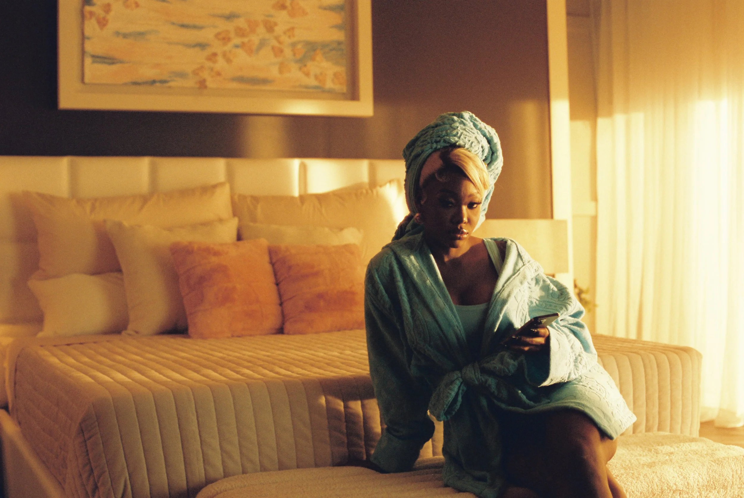A woman with a towel wrapped around her head and a robe on, sitting on a bed in a warmly lit hotel room, looking at her phone.