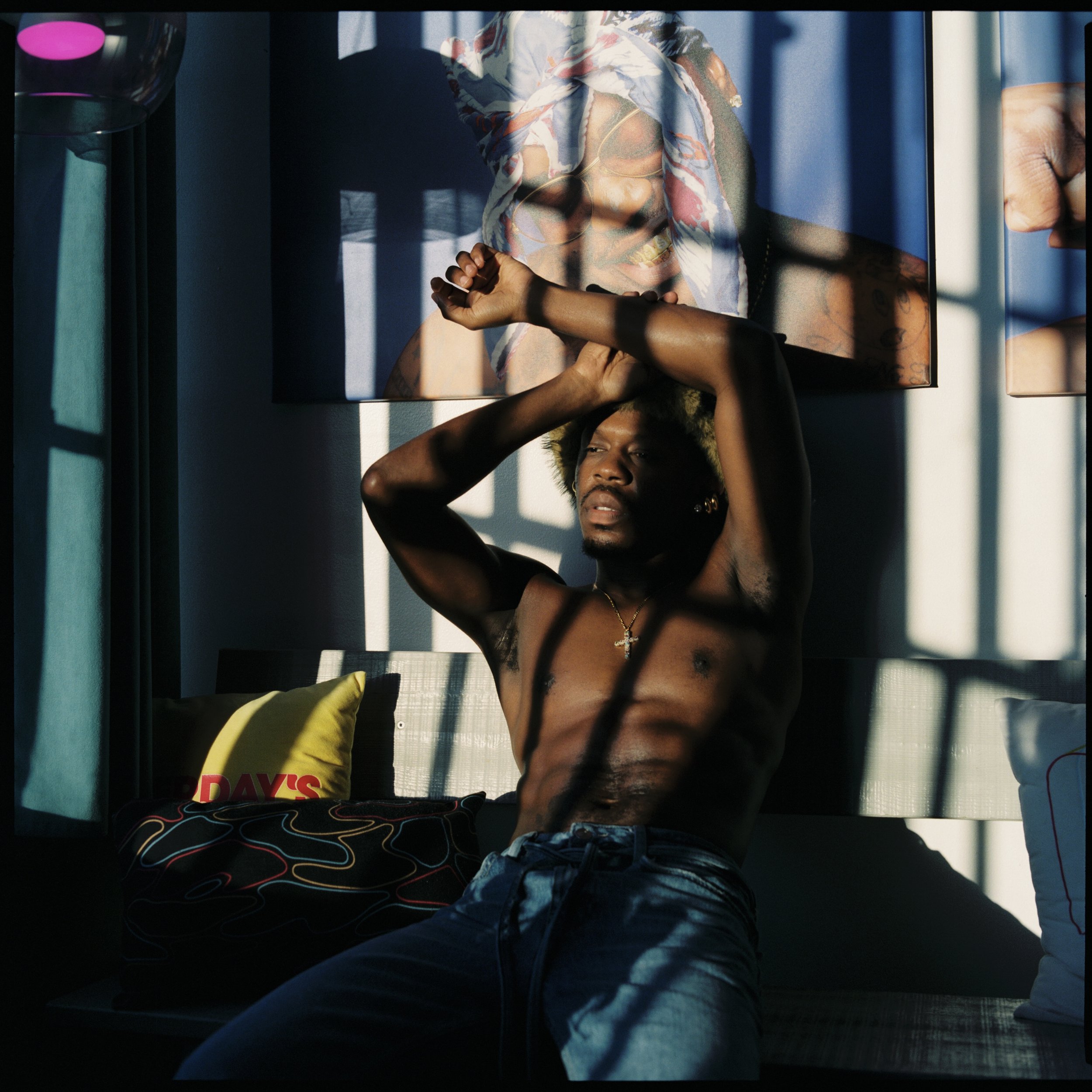 A shirtless man with curly hair and a necklace, sitting on a couch with sunlight casting shadows through window blinds, with a colorful portrait hanging on the wall behind him.