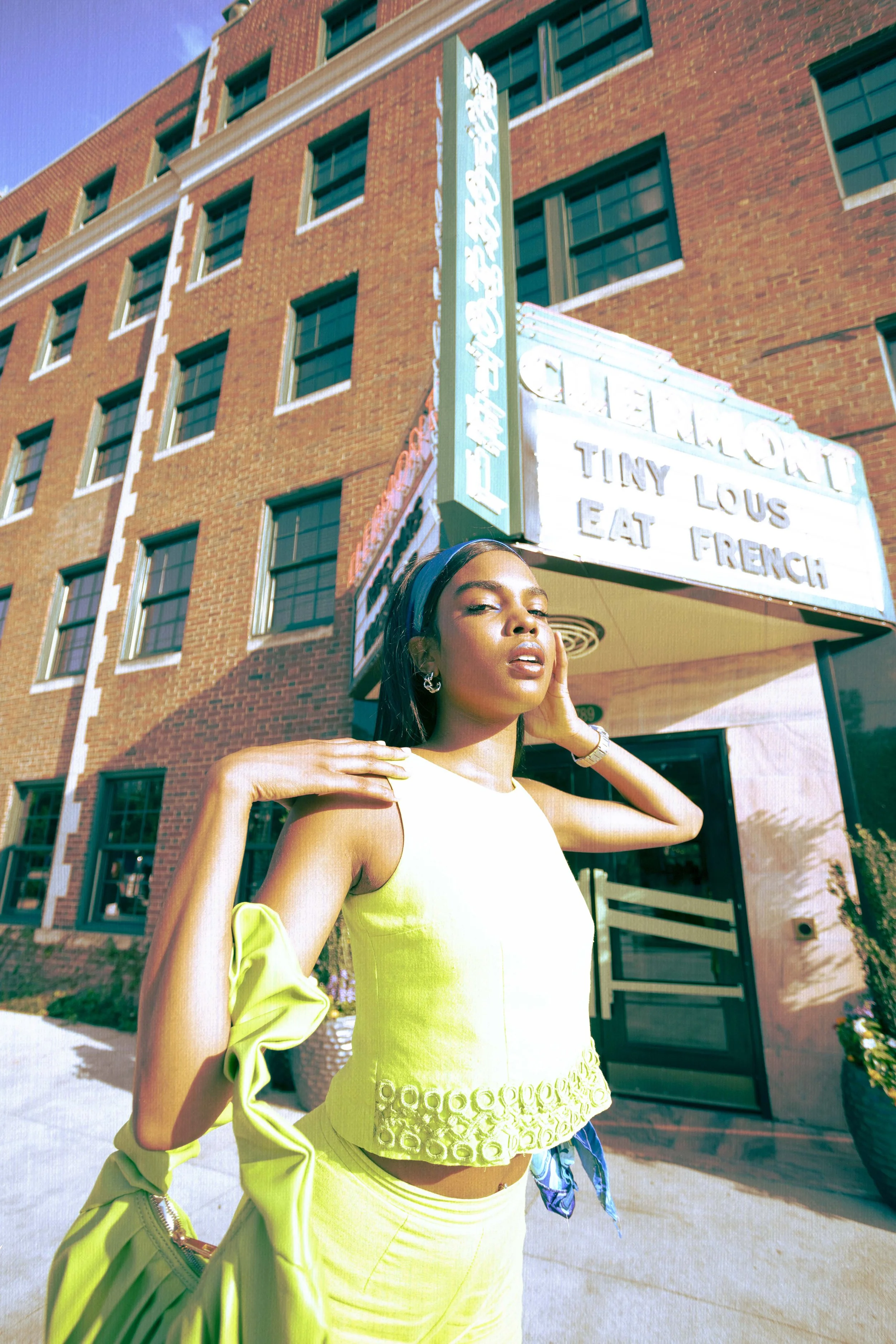 A woman in a yellow sleeveless top and matching skirt standing outside a brick building with a marquee sign. The sign reads "Tiny Lou's Eat French." She is touching her face and shoulder, with a slightly tilted head and confident expression, under a 