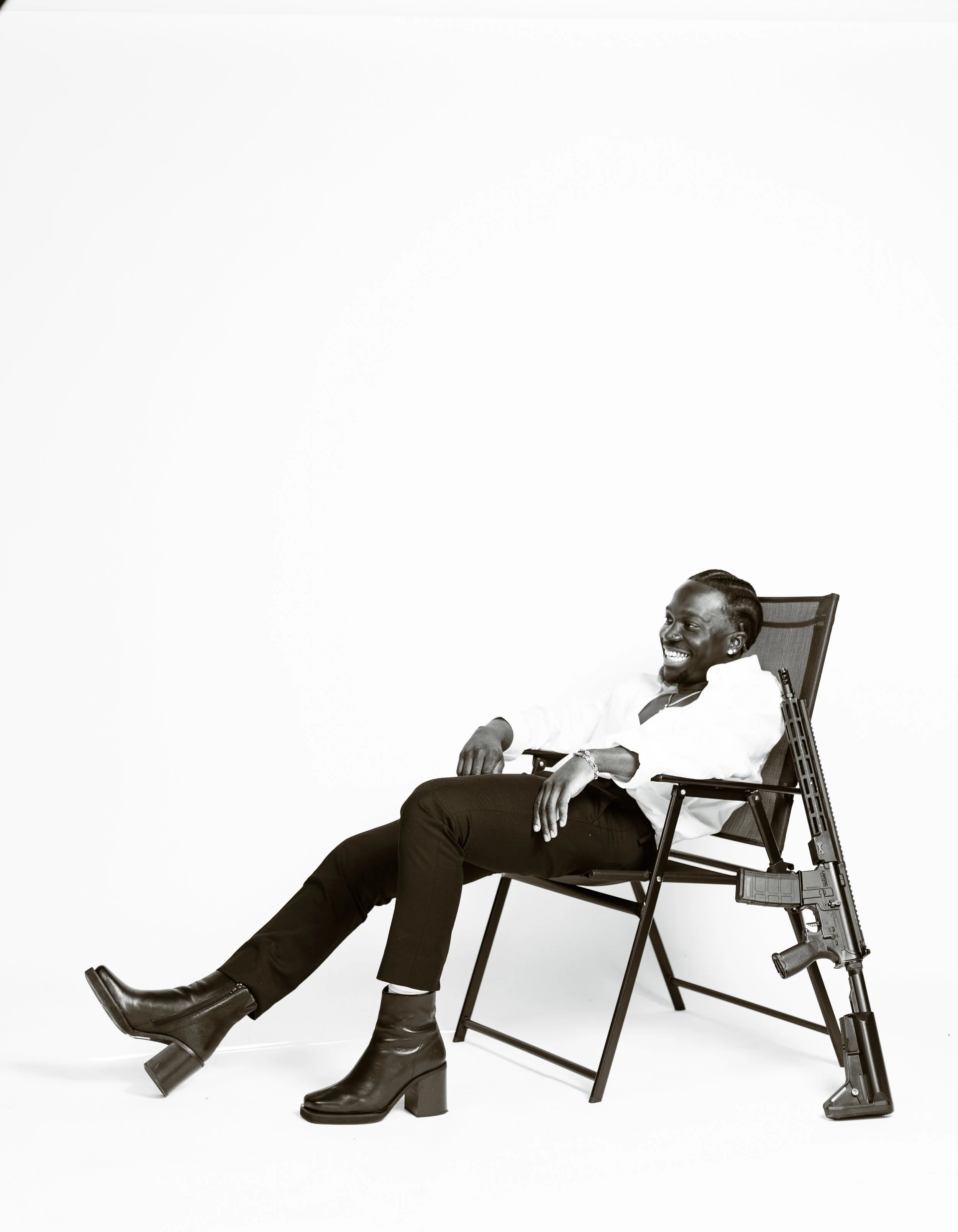 A man sitting in a lounge chair with a rifle leaning against the chair's side, smiling and appearing relaxed.