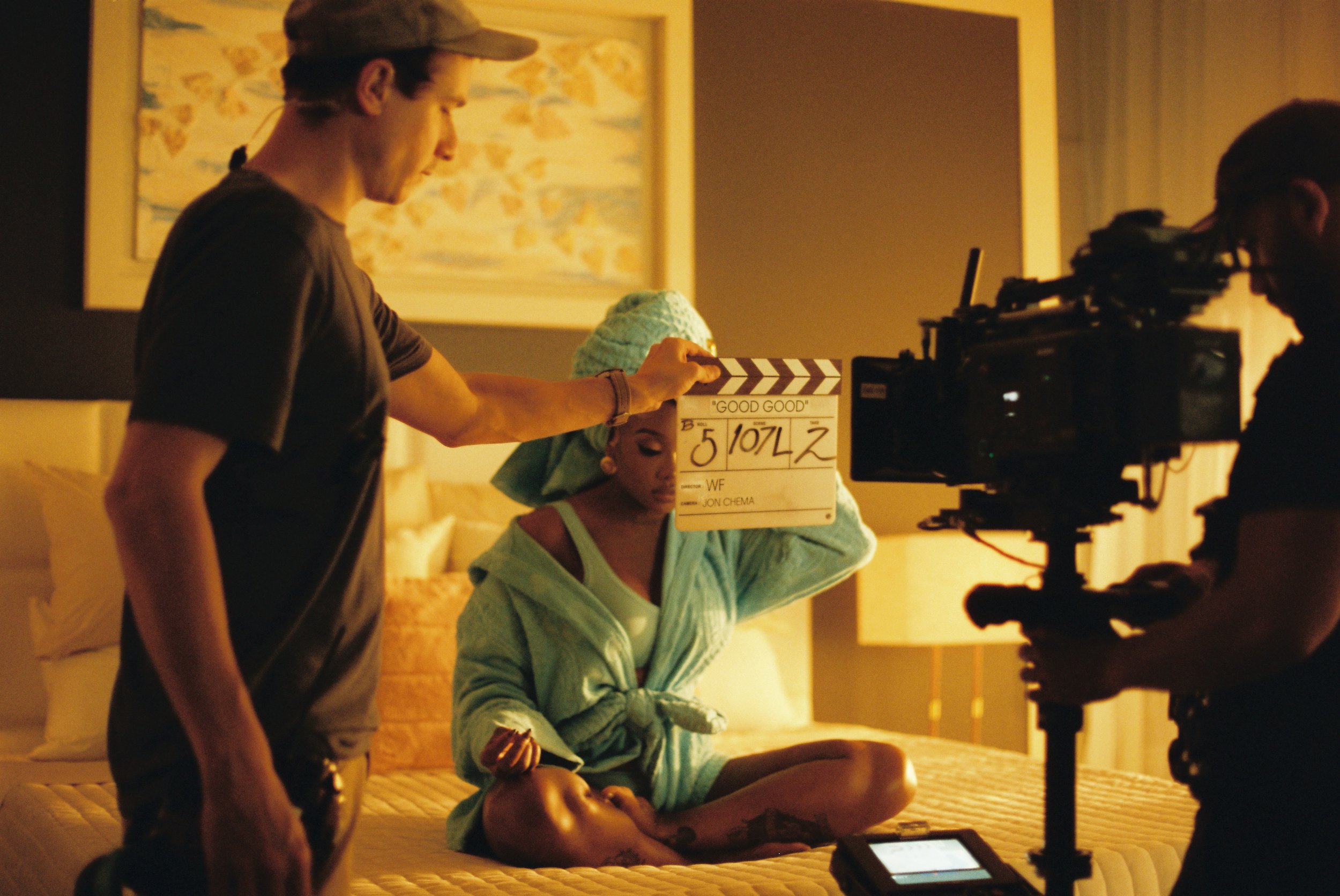 On a film set, a woman sits on a bed wearing a pastel green towel and hair wrap, holding a clapperboard, while a crew member adjusts it. A camera operator films her, in a warmly lit bedroom with artwork on the wall behind.