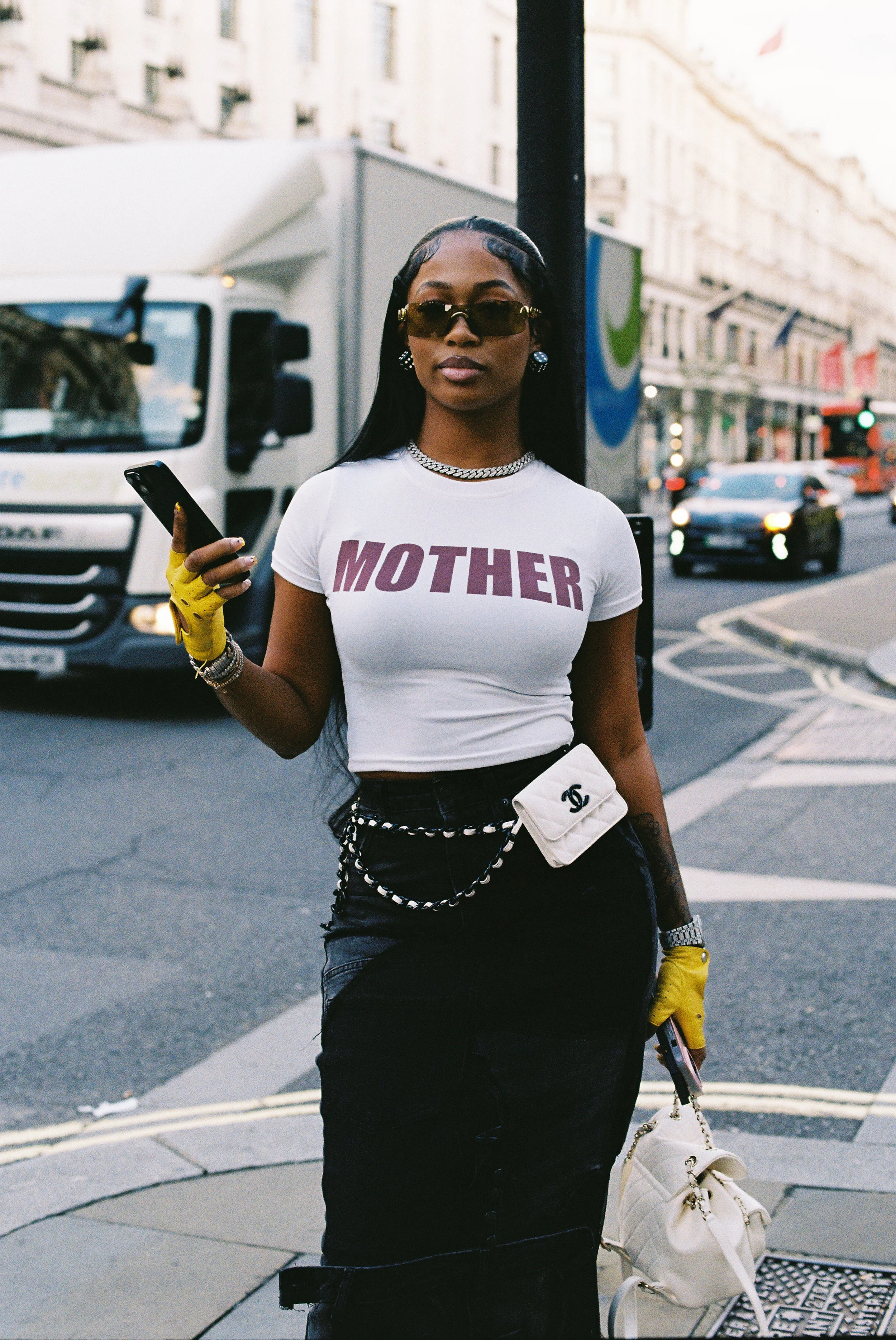 Stylish woman wearing sunglasses, a white T-shirt with 'MOTHER' written on it, black skirt, yellow gloves, and a white Chanel belt bag, holding a phone and a white handbag, standing on a city street.
