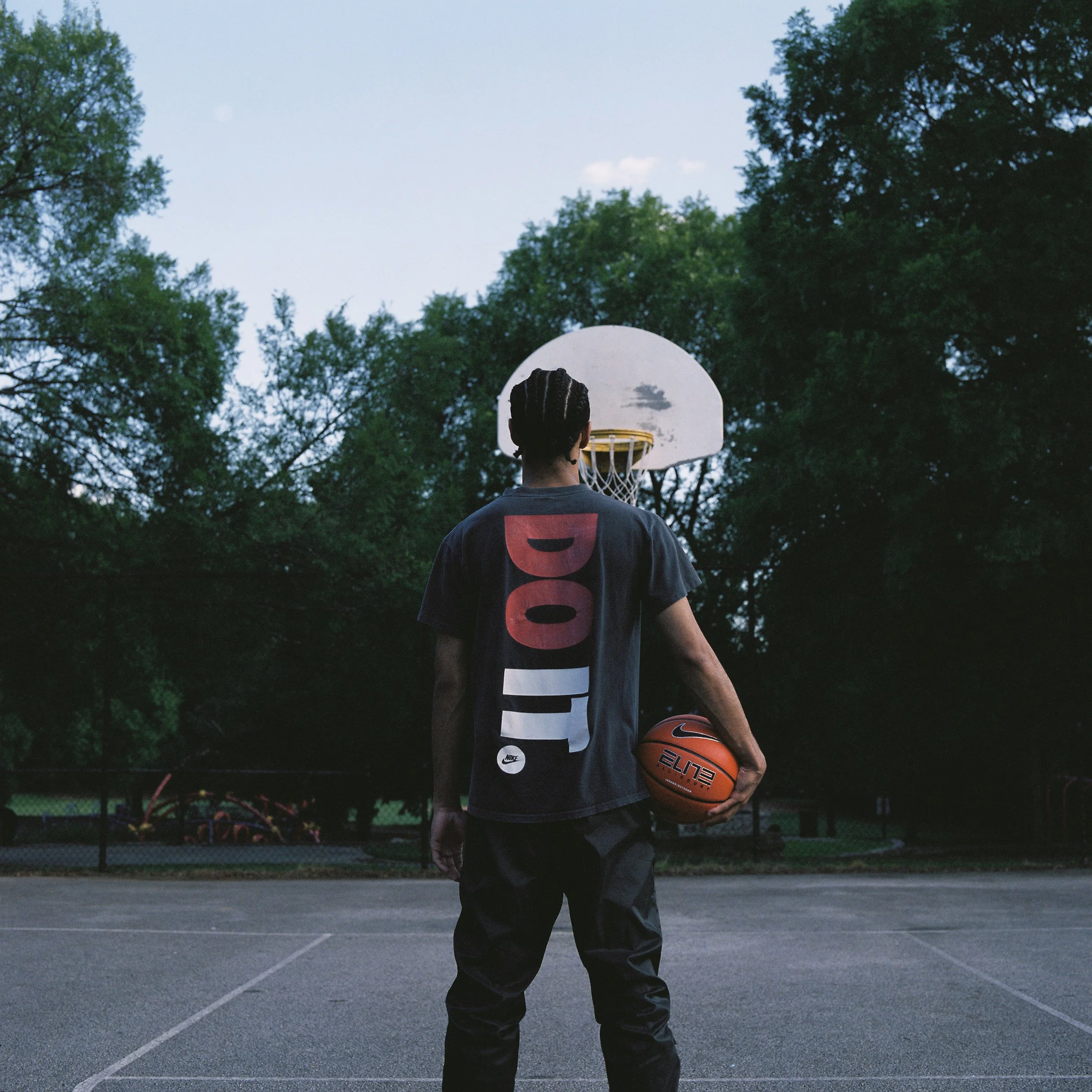 Back view of a person with braided hair standing on an outdoor basketball court during dusk, holding an orange basketball, facing a hoop surrounded by trees.