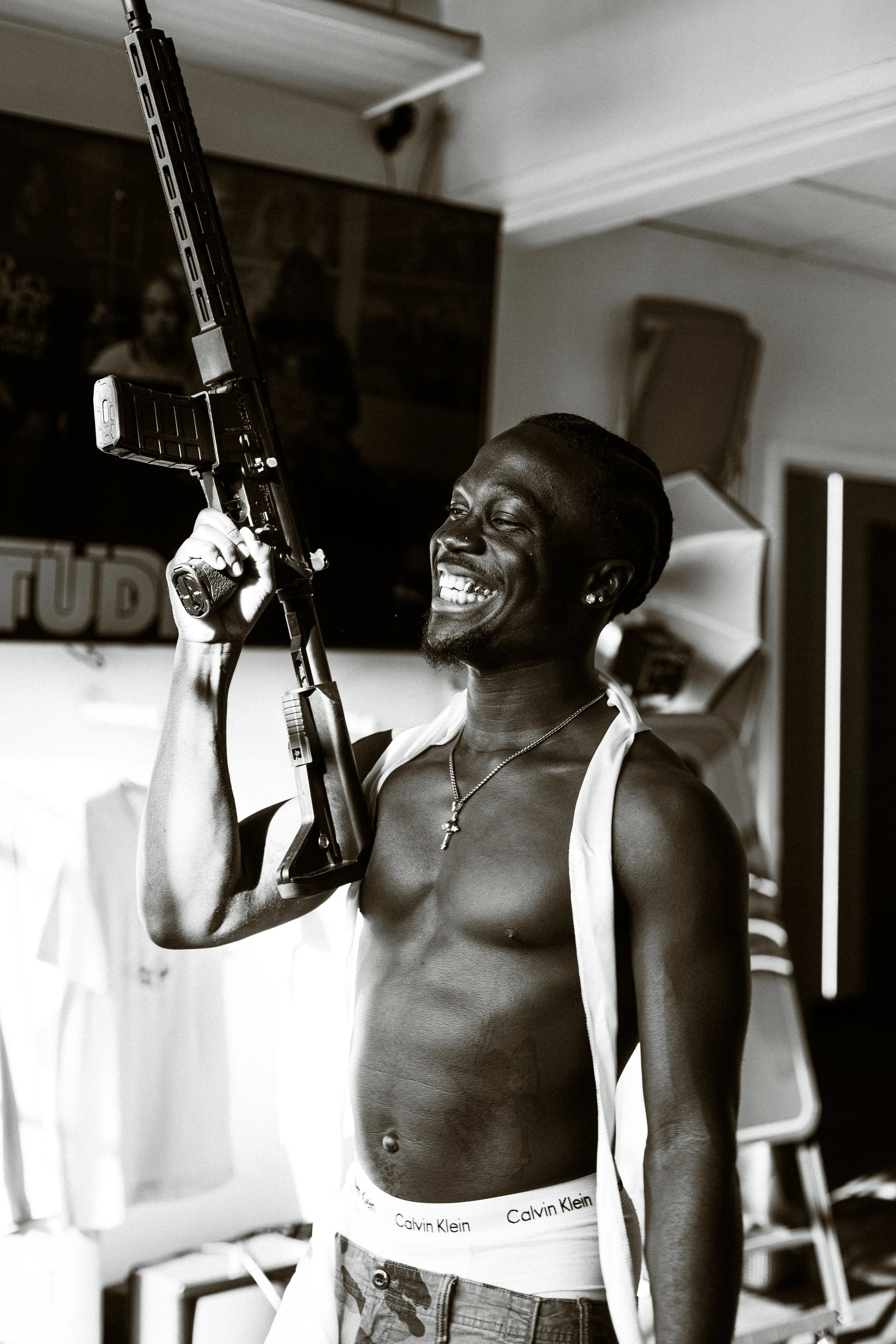 A smiling man holding a rifle indoors, shirtless with a white towel draped over his shoulders, wearing Calvin Klein underwear visible above denim shorts.