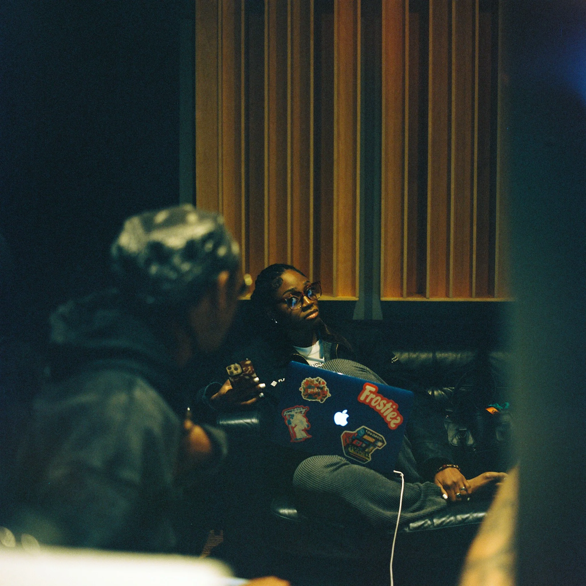 Two women sitting on a black leather couch in a studio or lounge setting. One woman, in the foreground, has light-colored hair, a hoodie, and glasses; she is holding a smartphone. The other woman, in the background, has braided hair, glasses, and is 