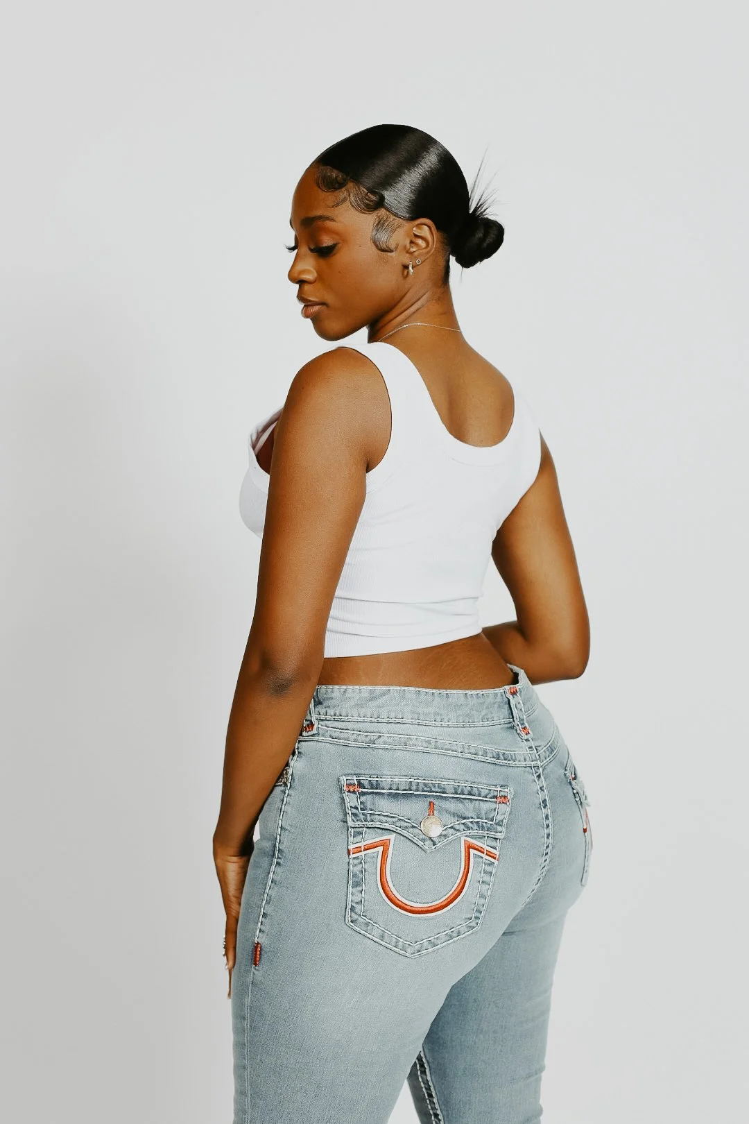 A woman with dark hair styled in a low bun, wearing a white sleeveless crop top and light blue jeans with a decorative back pocket, standing against a plain white background.
