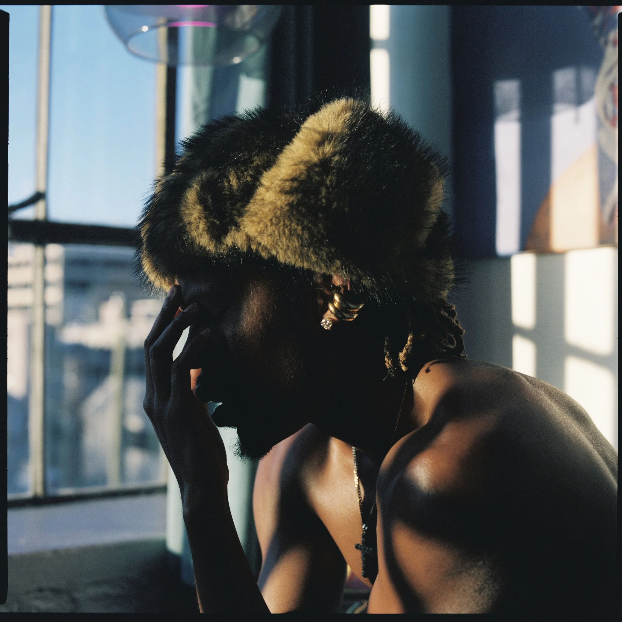 Side profile of a shirtless man wearing a fur hat, with a hand covering part of his face, in an indoor setting with sunlight and windows in the background.