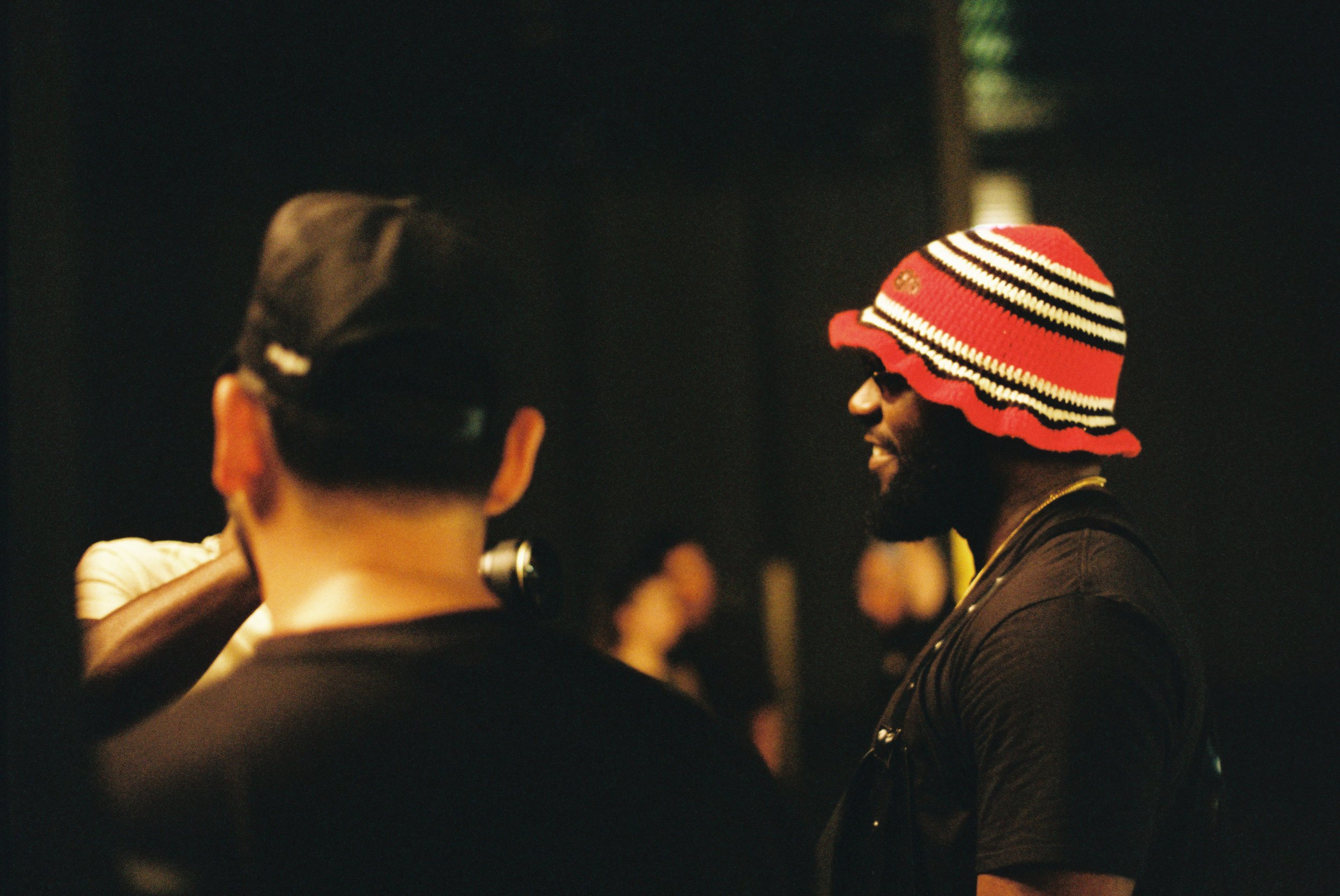 A man wearing a colorful striped hat and sunglasses, smiling, appears to be engaged in a conversation with others in a dimly lit setting.