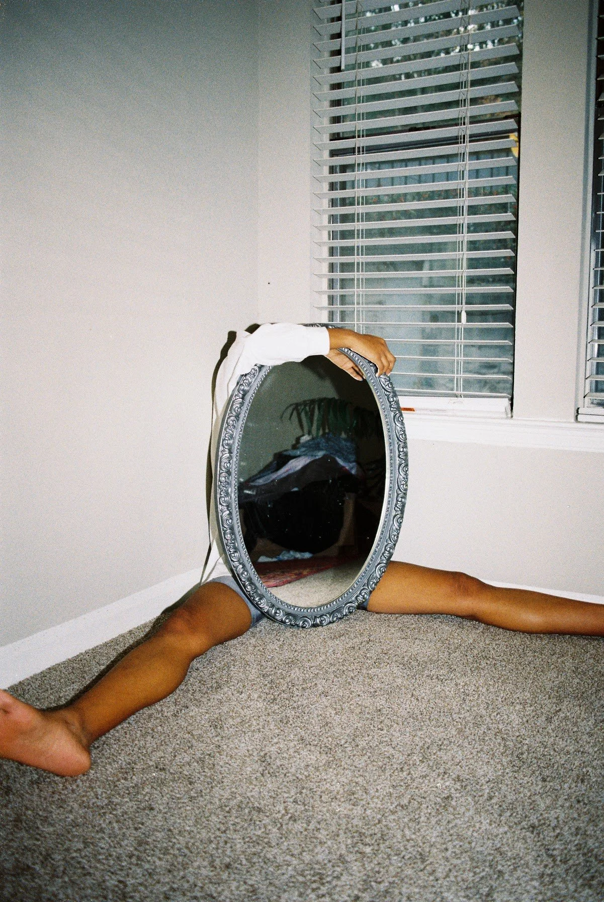 Person's legs and hand reflected in a decorative oval mirror placed on the carpeted floor, with a window and blinds in the background.
