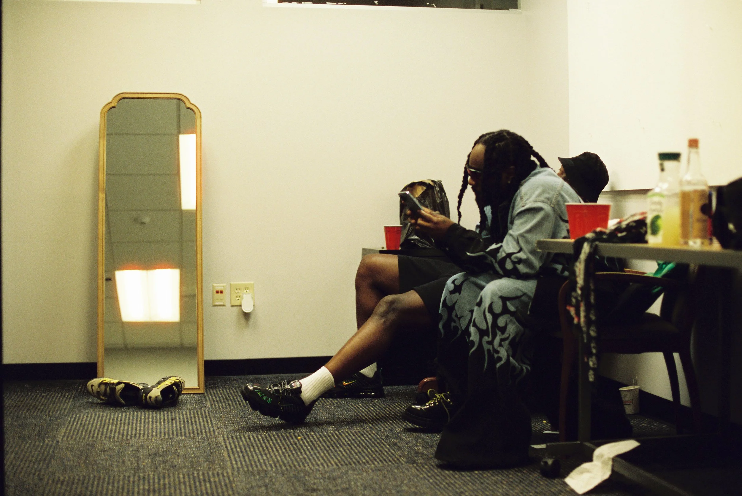 Two people sitting against a wall in a crowded room, looking at a phone, with shoes, bottles, and red cups around them.