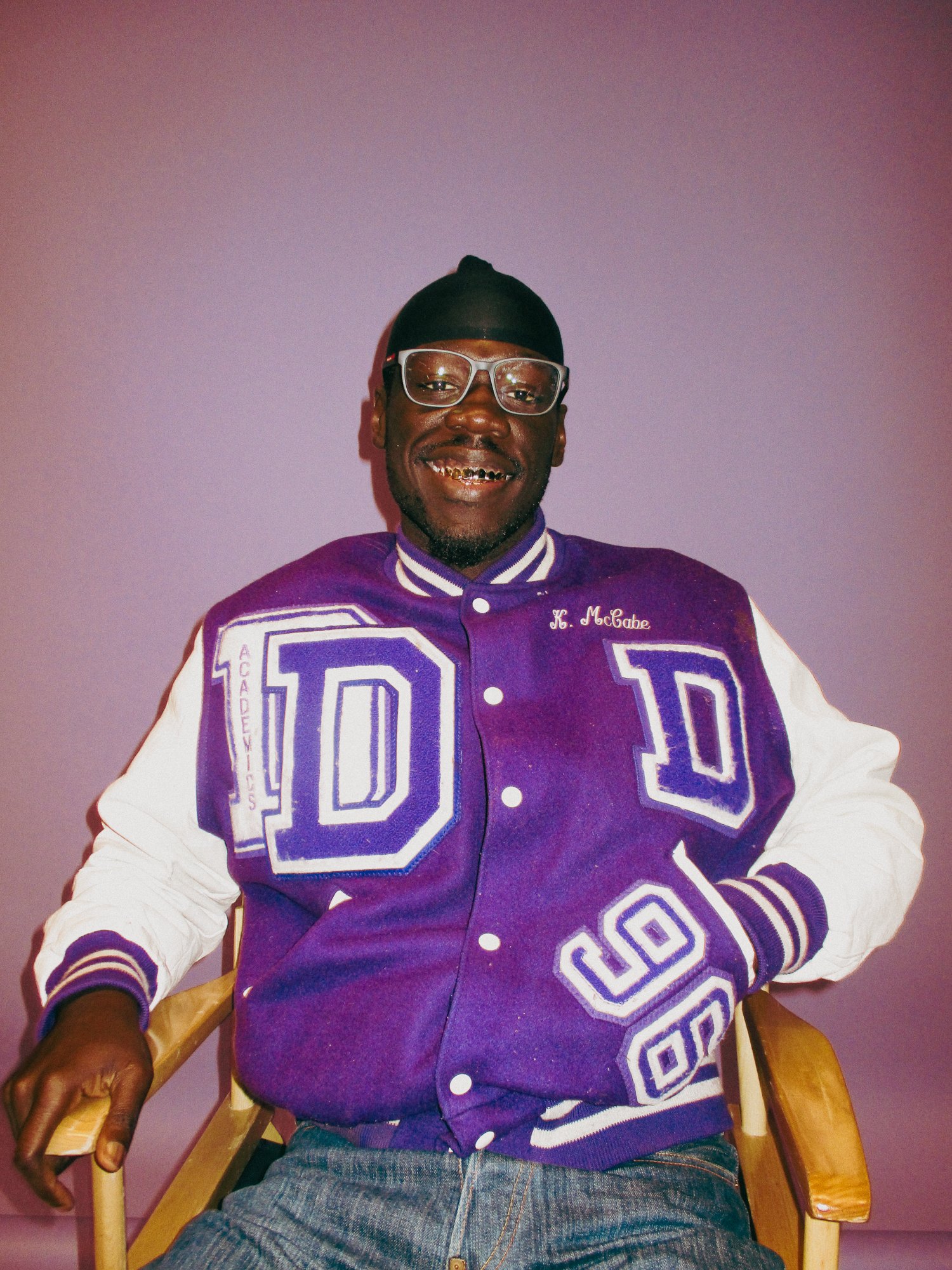 A smiling man wearing a purple and white varsity jacket with 'D' patches, glasses, and a black durag, sitting on a wooden chair against a plain wall.