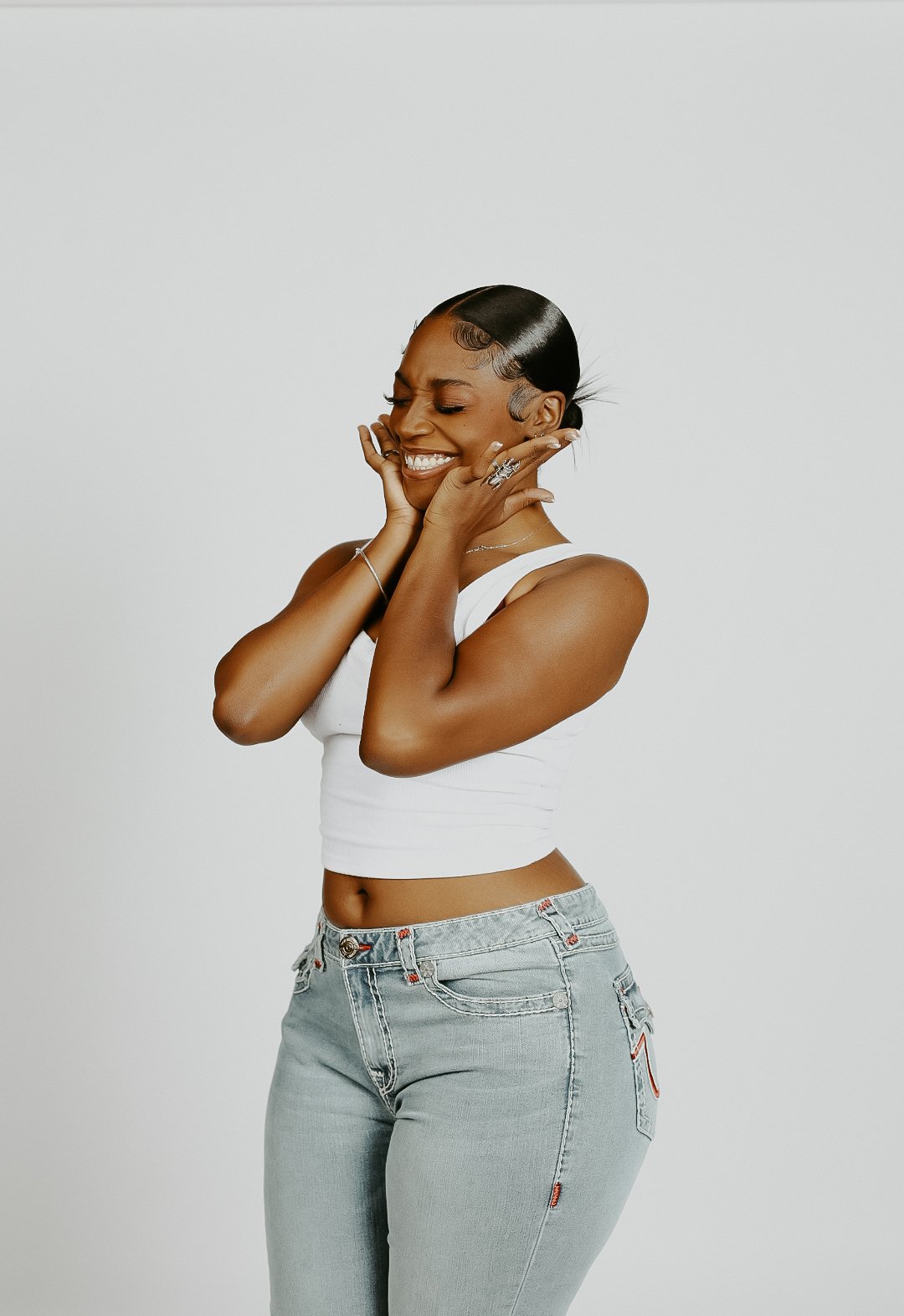 Young woman with short black hair smiling with hands on her face, wearing a white sleeveless top and light gray jeans against a plain white background.