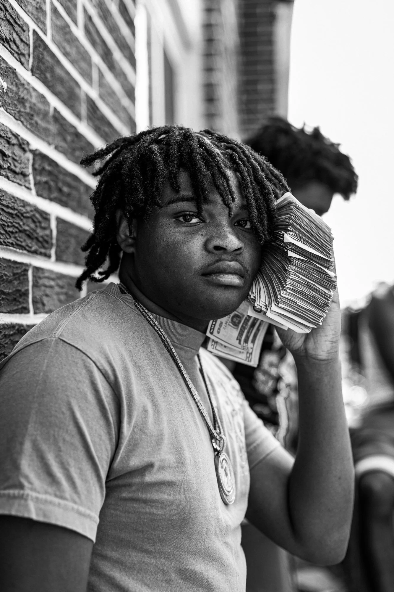 Young man with dreadlocks holding a large stack of cash to his ear, standing against a brick wall in black and white.