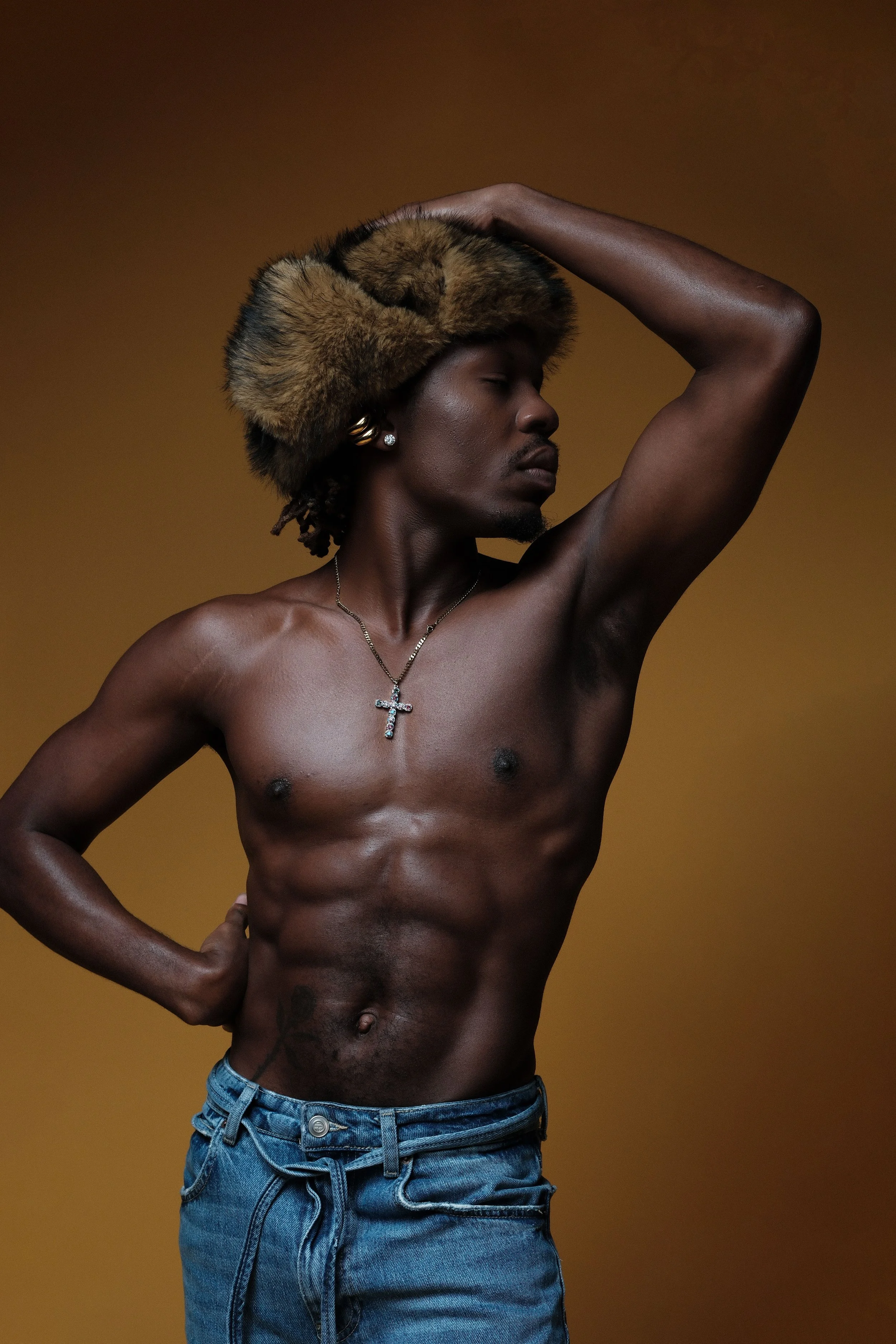 A shirtless young man with dark skin, wearing a brown fur hat, blue jeans, and jewelry including a necklace with a cross and earrings, posing with one arm raised and the other hand on his hip against a brown background.