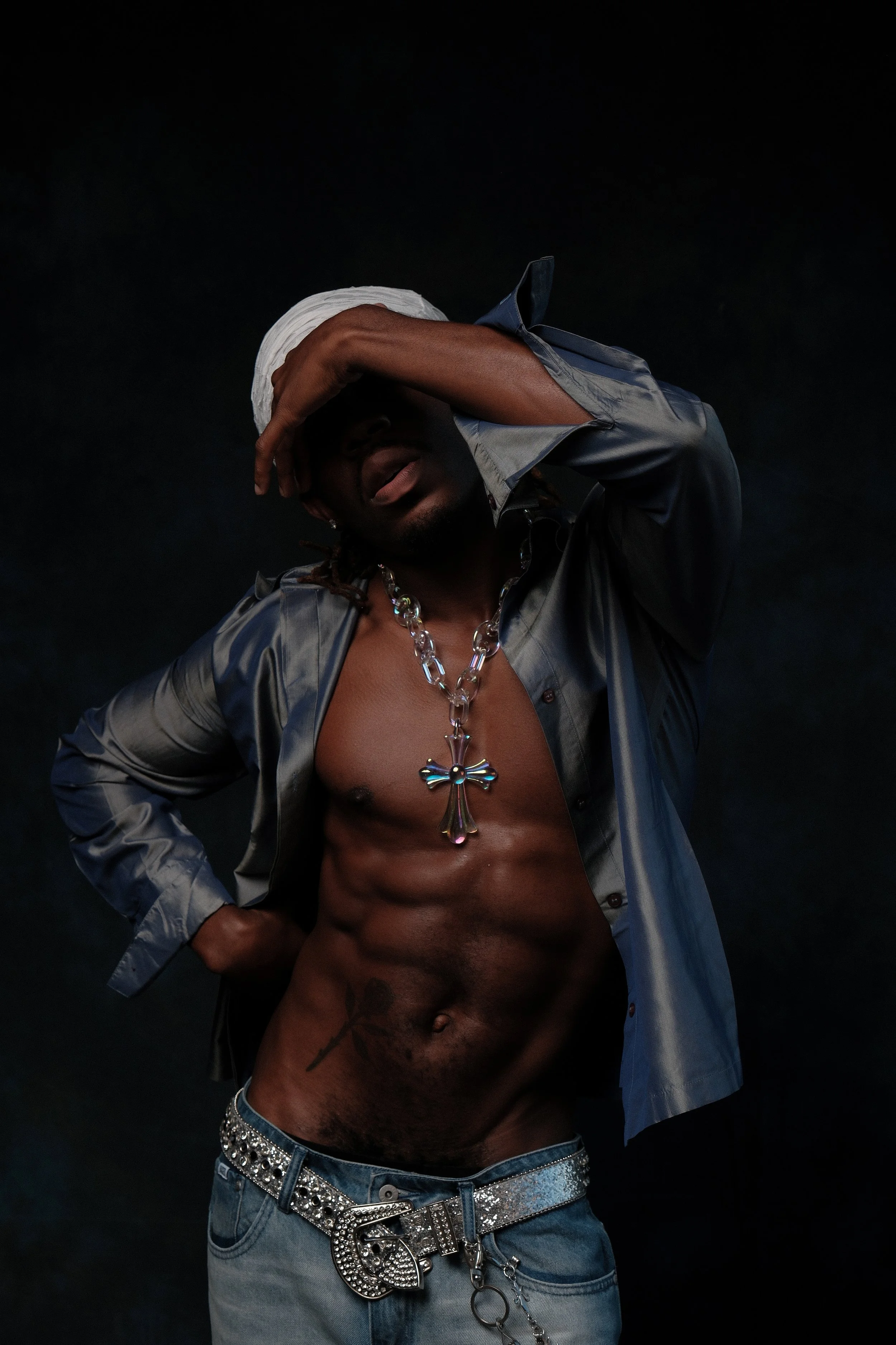 A shirtless man wearing jeans, a large cross necklace, and a shiny belt with jewelry. He has a white cloth on his head and covers part of his face with his hand, posing against a dark background.
