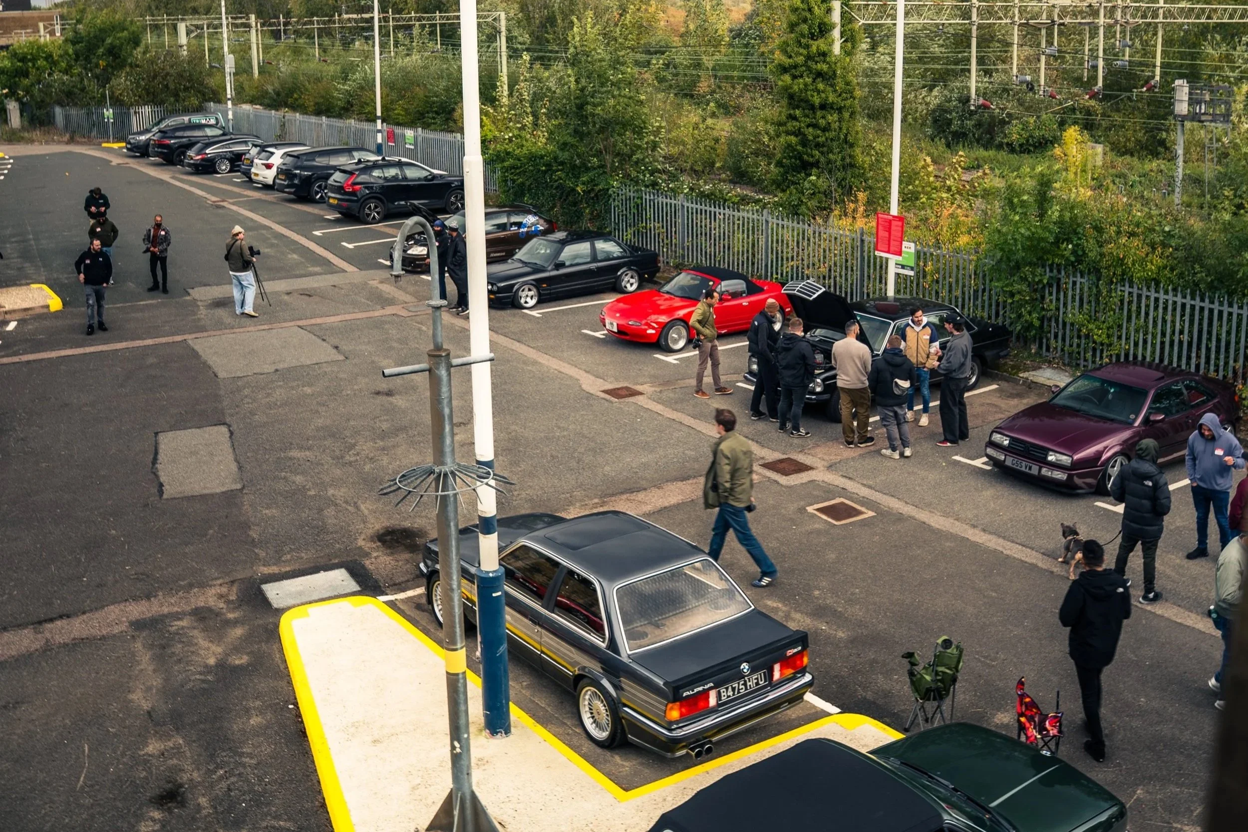 Black Porsche 911 with custom wheels parked under an overpass, with a yellow and red Mazda RX-7 beside it, at what appears to be a car meet, with people walking around and city buildings in the background.