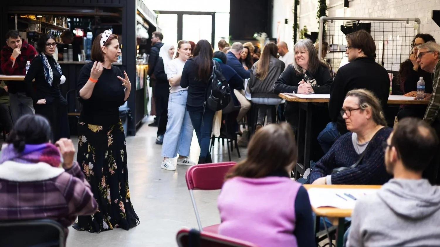 A group of people gathered in a lively indoor space with some standing in line, sitting, and engaging in conversation, possibly at a social event or workshop. There is a bar or counter in the background.